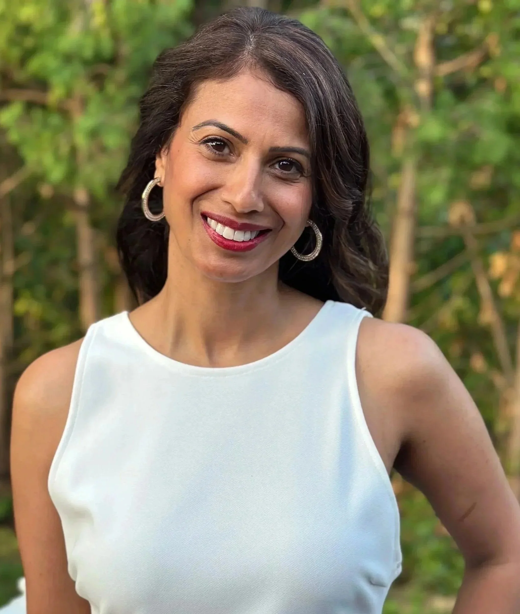 Smiling woman with dark brown hair, wearing hoop earrings and a sleeveless white top, standing outdoors with green foliage in the background.