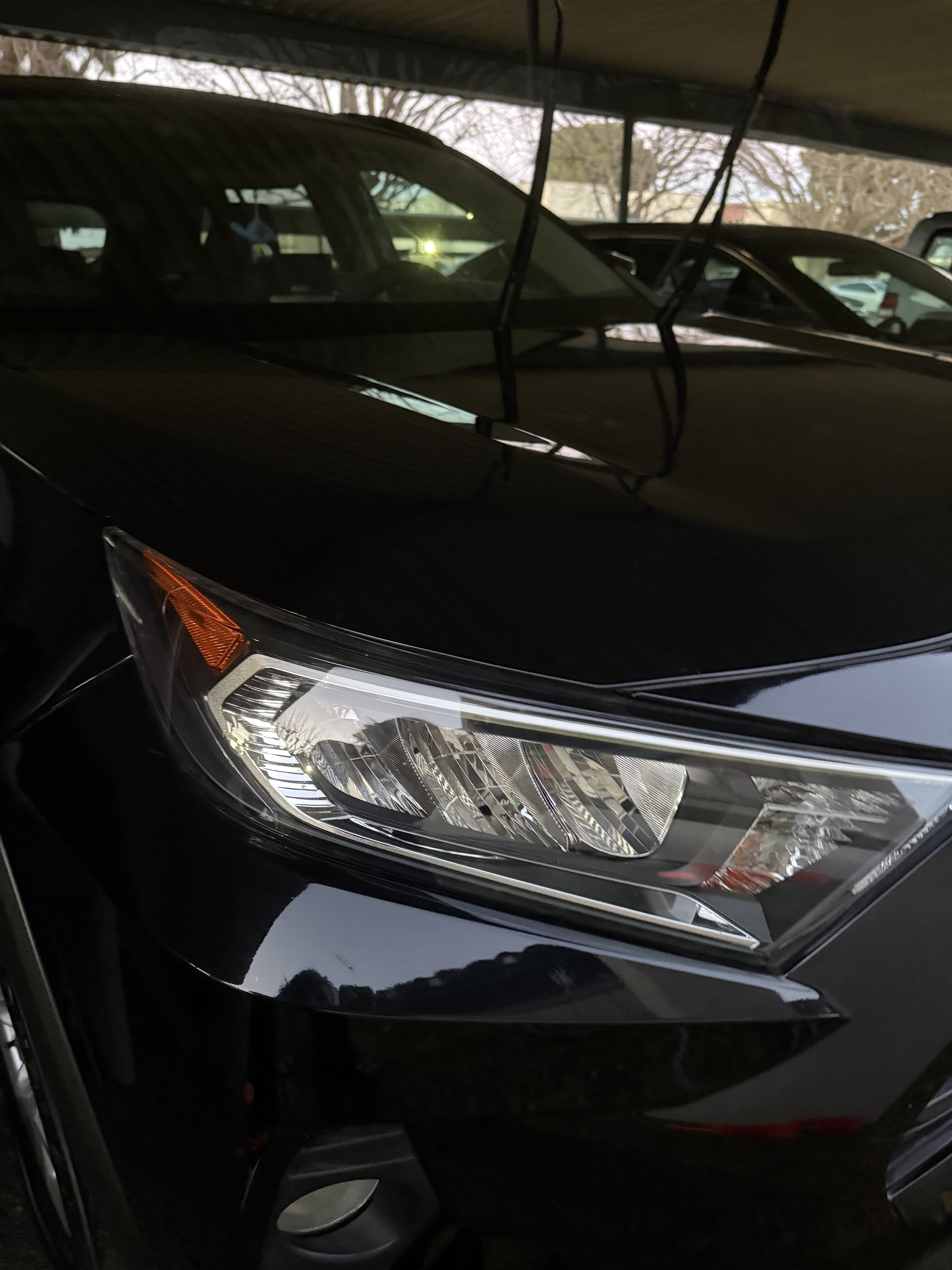 Close-up of a black car's front headlight and part of the hood in a dimly lit parking garage.