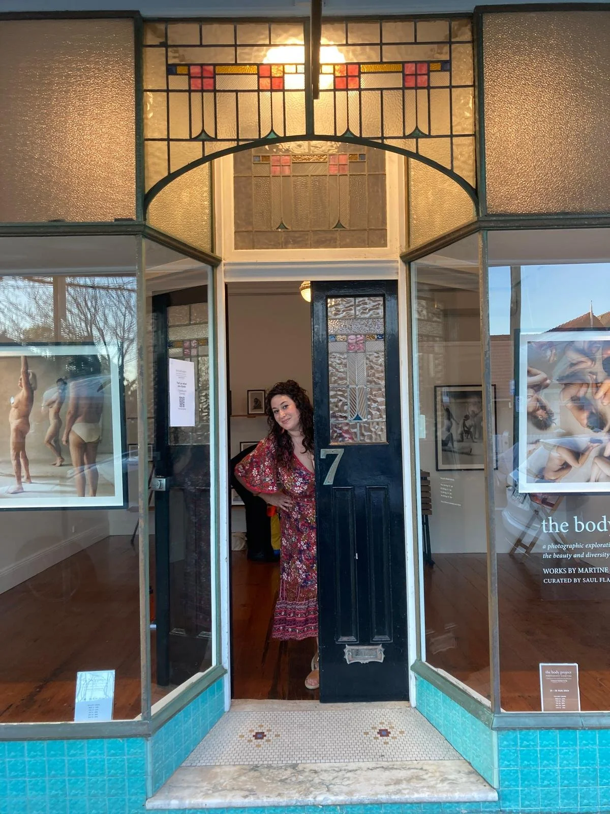 A woman with curly hair, wearing a floral dress, standing partially inside a gallery or store called "the body project," peeking out from behind a black door with the number 7, with art and photographs visible inside, and stained glass window details above the doorway.
