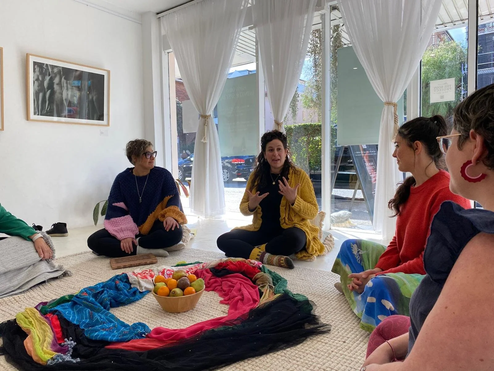 Group of women sitting on the floor in a circle in a cozy room with large windows, curtains, and framed photos on the wall, engaged in a discussion or activity about art and color.
