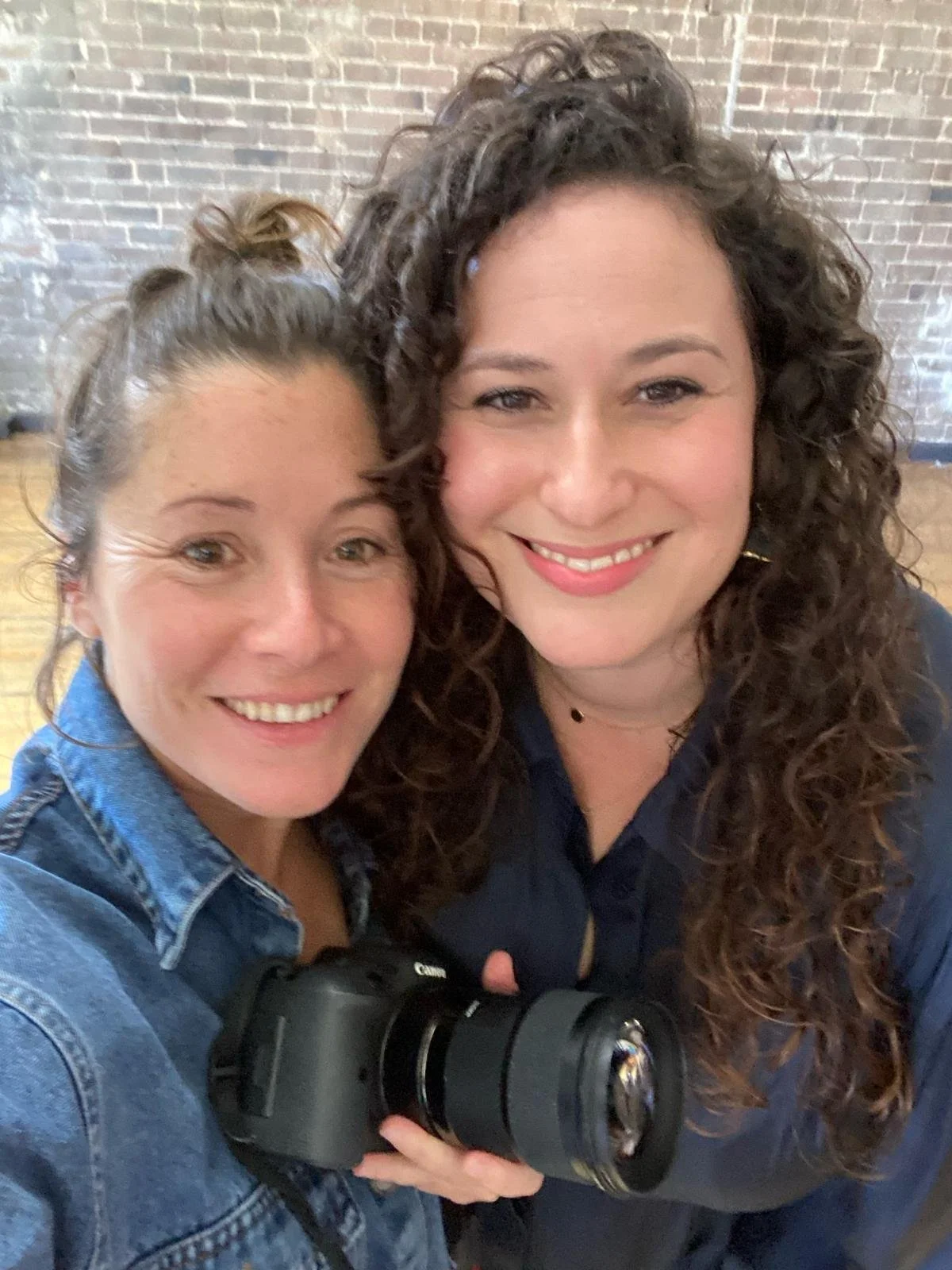 Two women smiling and taking a selfie, one holding a camera, with a brick wall in the background.
