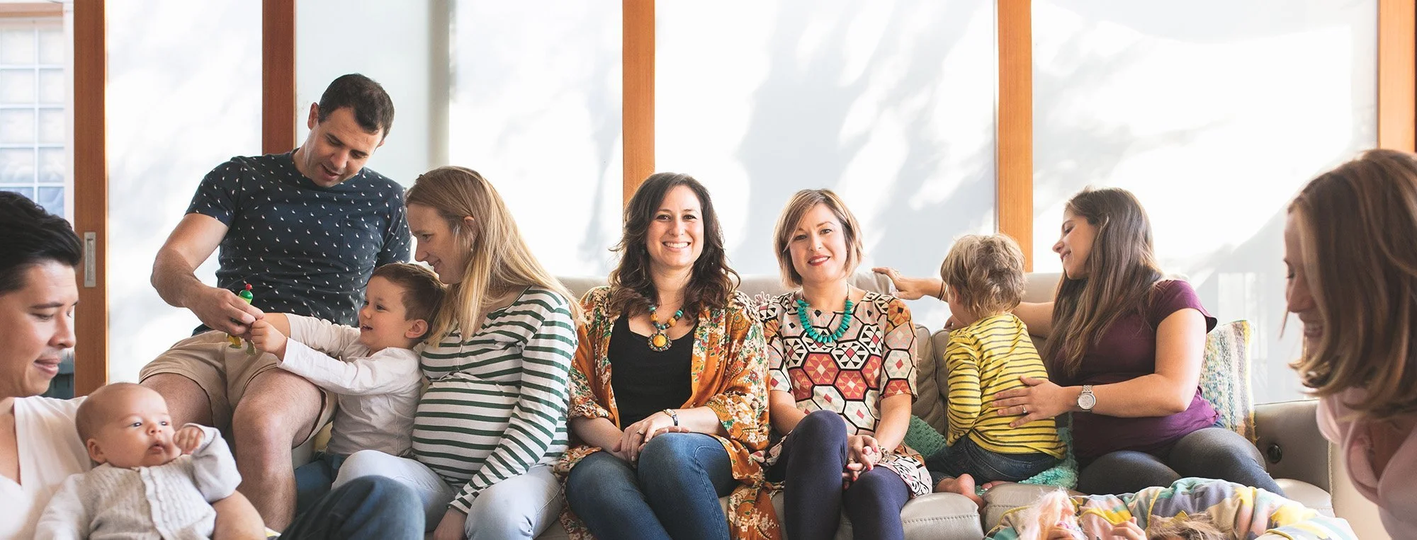 Group of adults and children sitting on a couch and smiling during a gathering in a bright room with large windows.