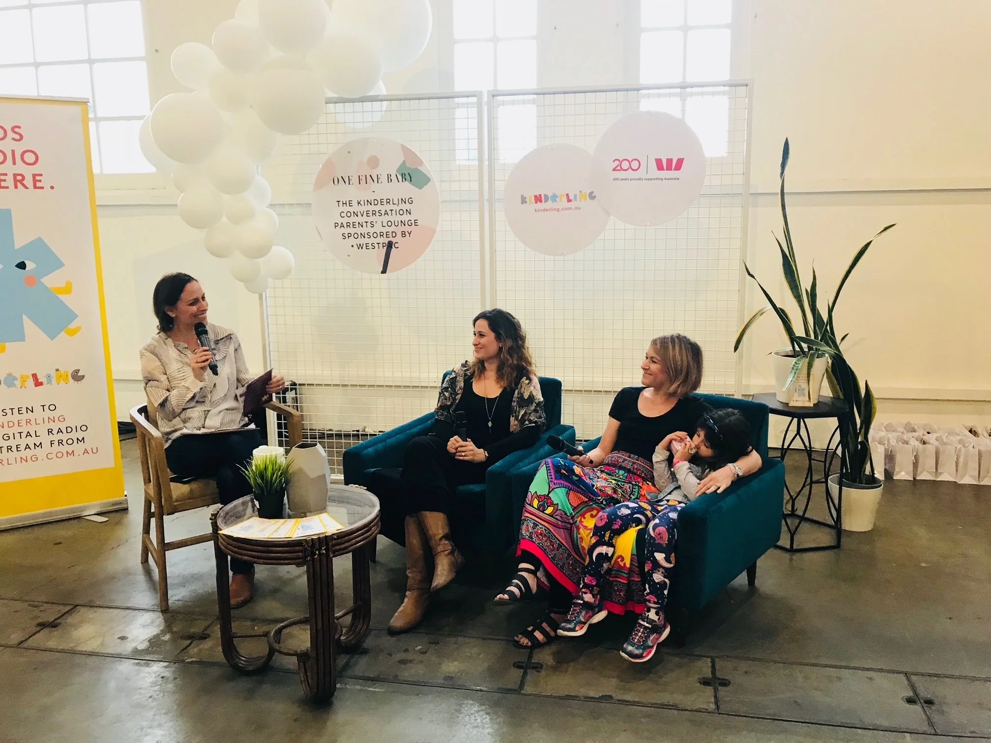 Three women sitting on a teal sofa in a bright room with large windows, participating in a panel discussion. One woman is speaking into a microphone, while the other two listen. There are white balloons, a white grid backdrop with logos, potted plants, and a yellow sign with event information.