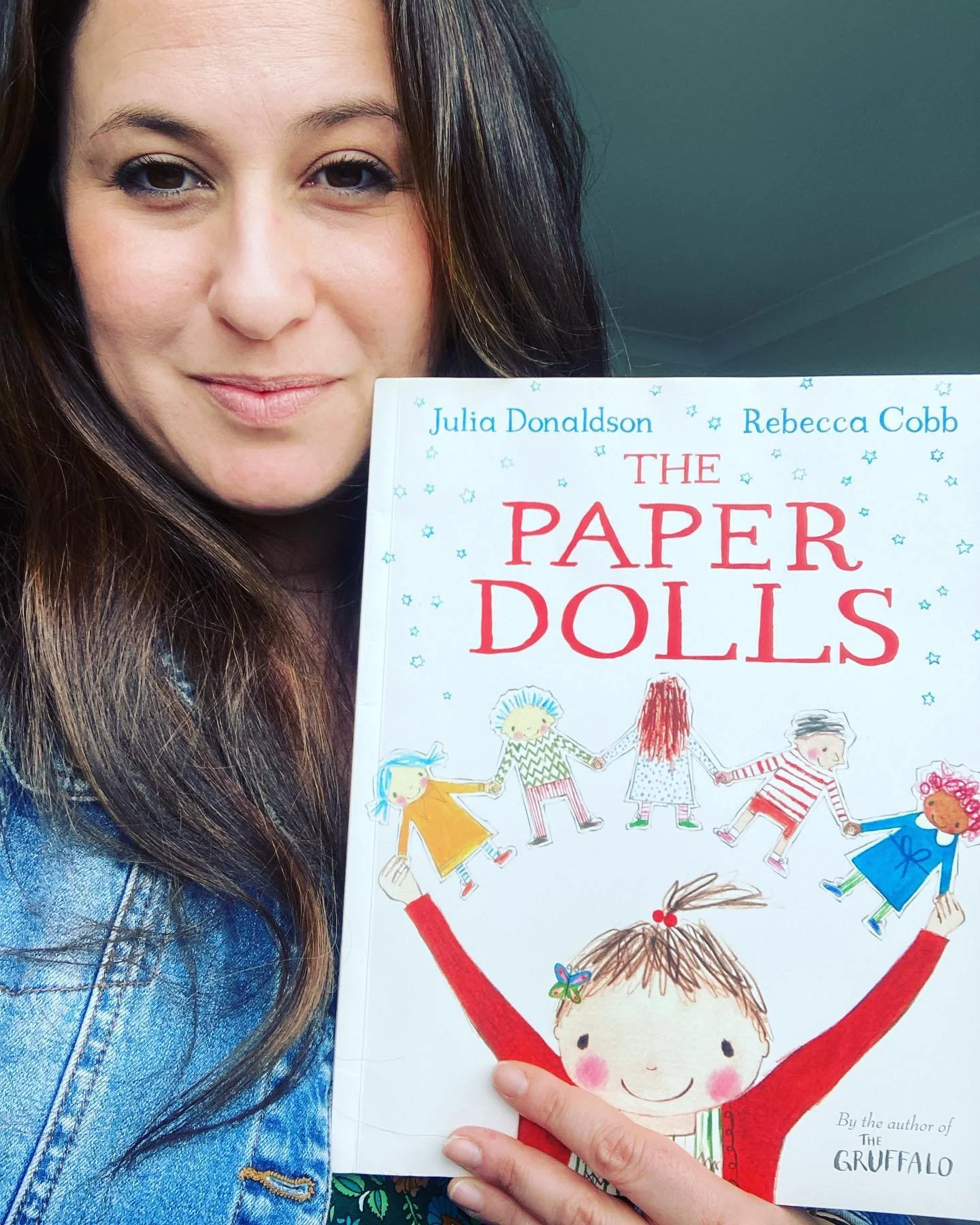 A woman with long dark hair holding a children's book titled 'The Paper Dolls' by Julia Donaldson and Rebecca Cobb. She is smiling slightly and is wearing a denim jacket. The book cover features an illustration of a girl with rosy cheeks and a butterfly in her hair, holding paper dolls.