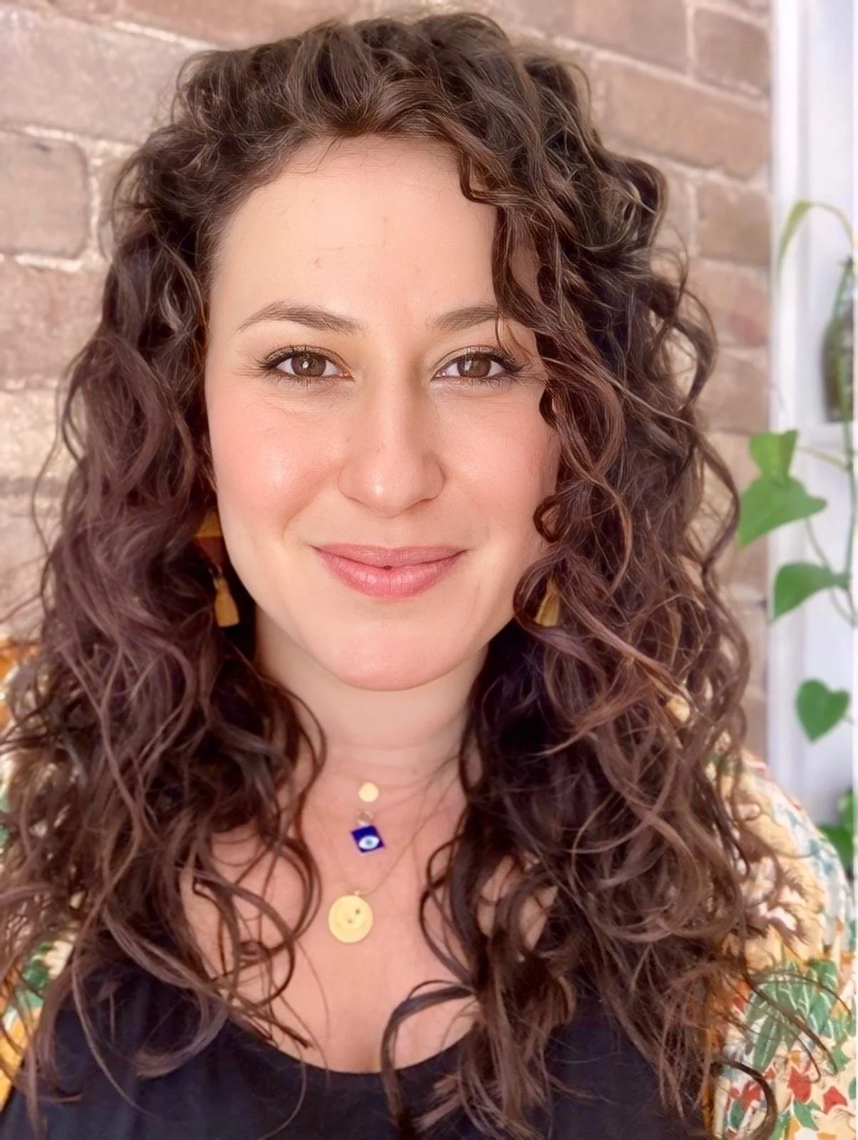 A woman with long, curly brown hair smiling, wearing a black top and layered necklaces, standing against a brick wall with some green plants in the background.