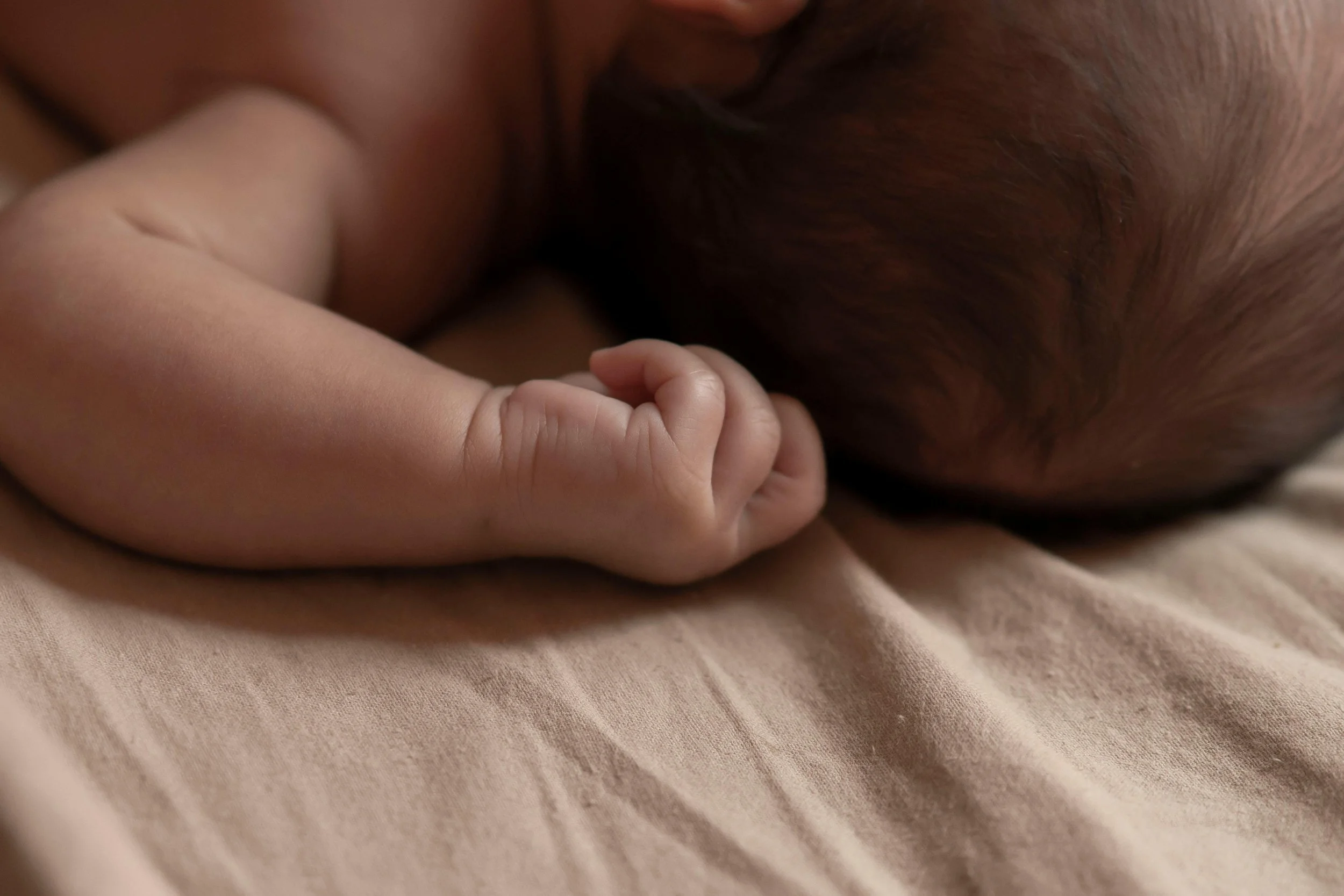 Close-up of a newborn baby lying on a soft surface, showing tiny hand and head with hair.