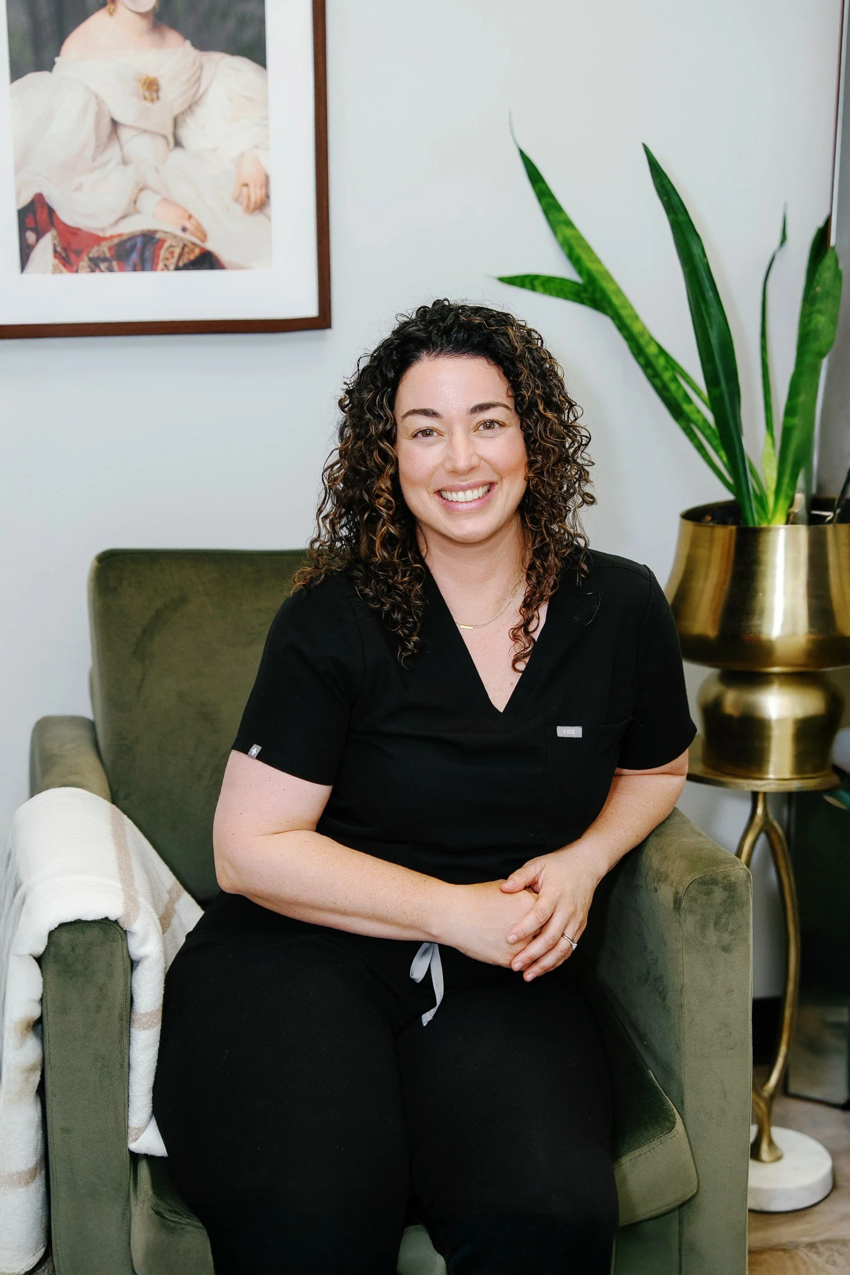 A woman with curly hair smiling while sitting on a green armchair in an indoor space. There is a large potted plant in a gold-colored planter to her right and a framed portrait hanging on the wall behind her.