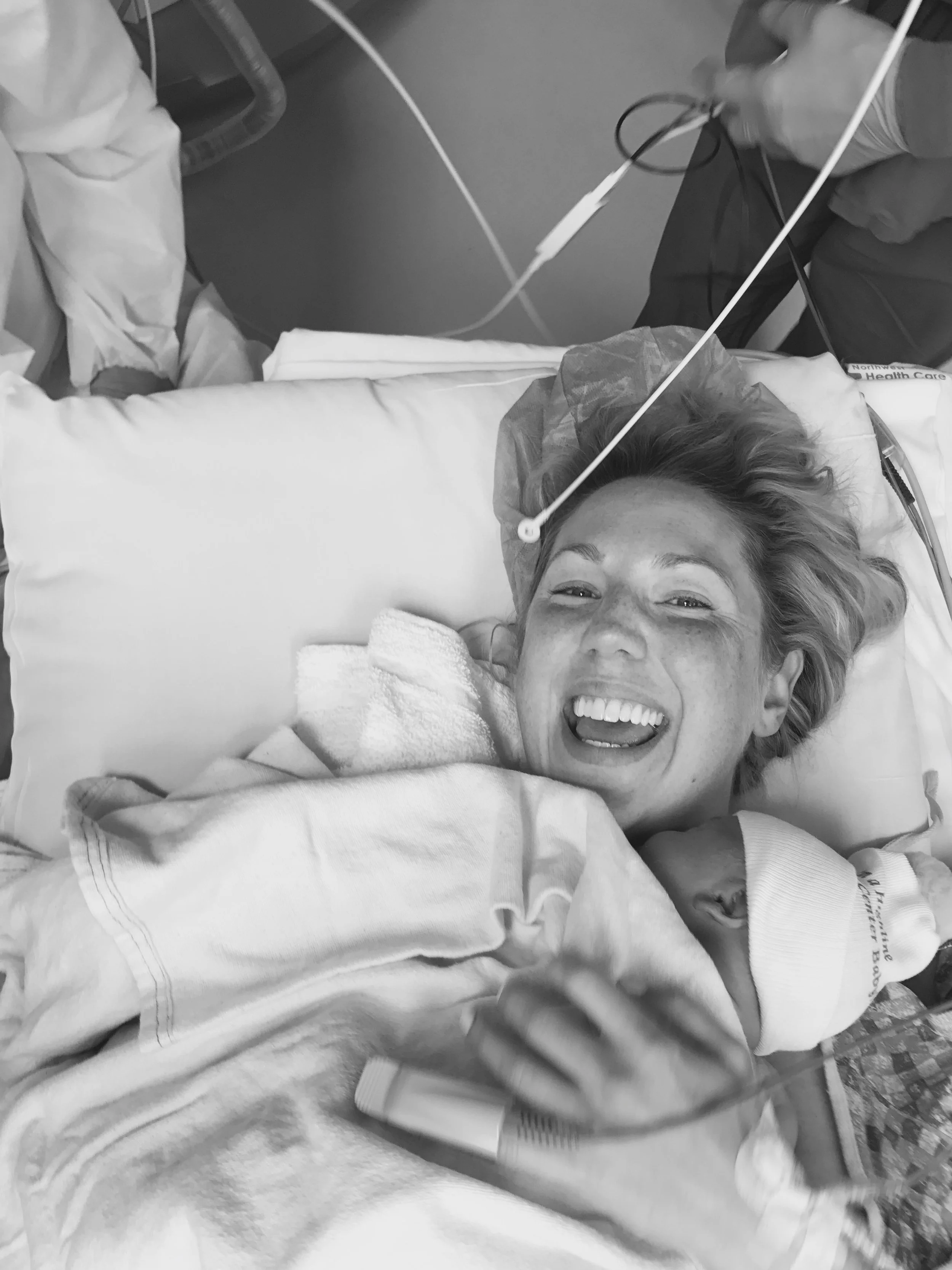 A woman lying in a hospital bed, smiling enthusiastically, with a newborn baby she is holding.