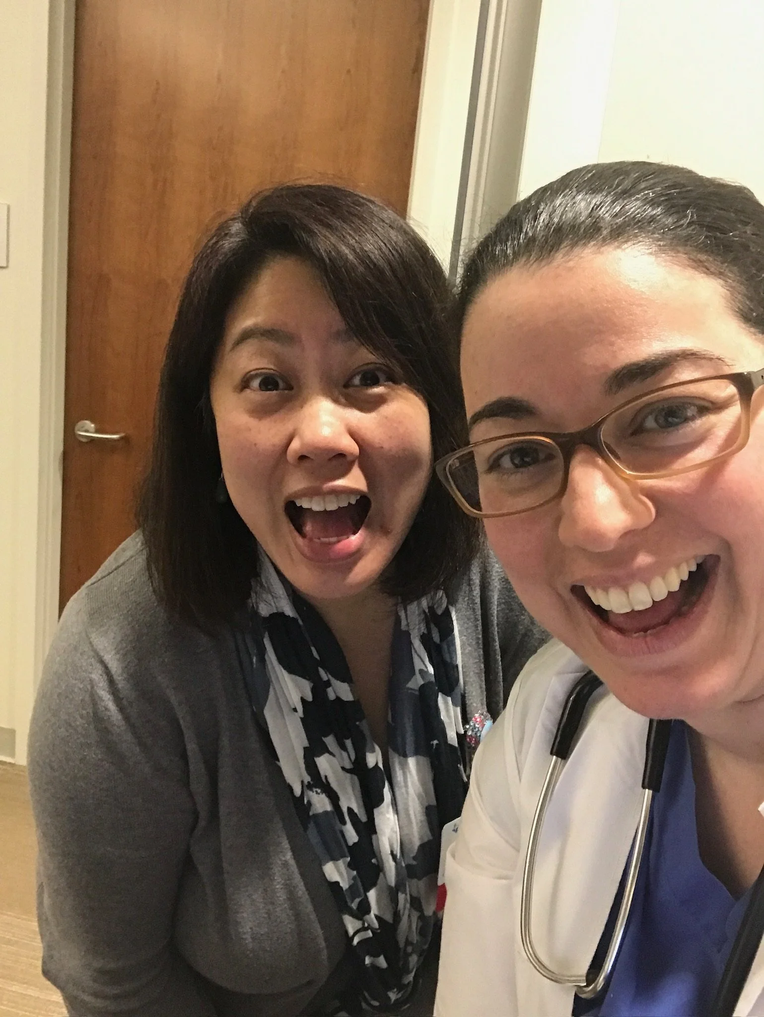 Two women smiling and taking a selfie together indoors, one wearing glasses and a stethoscope, the other with a patterned scarf and gray cardigan.