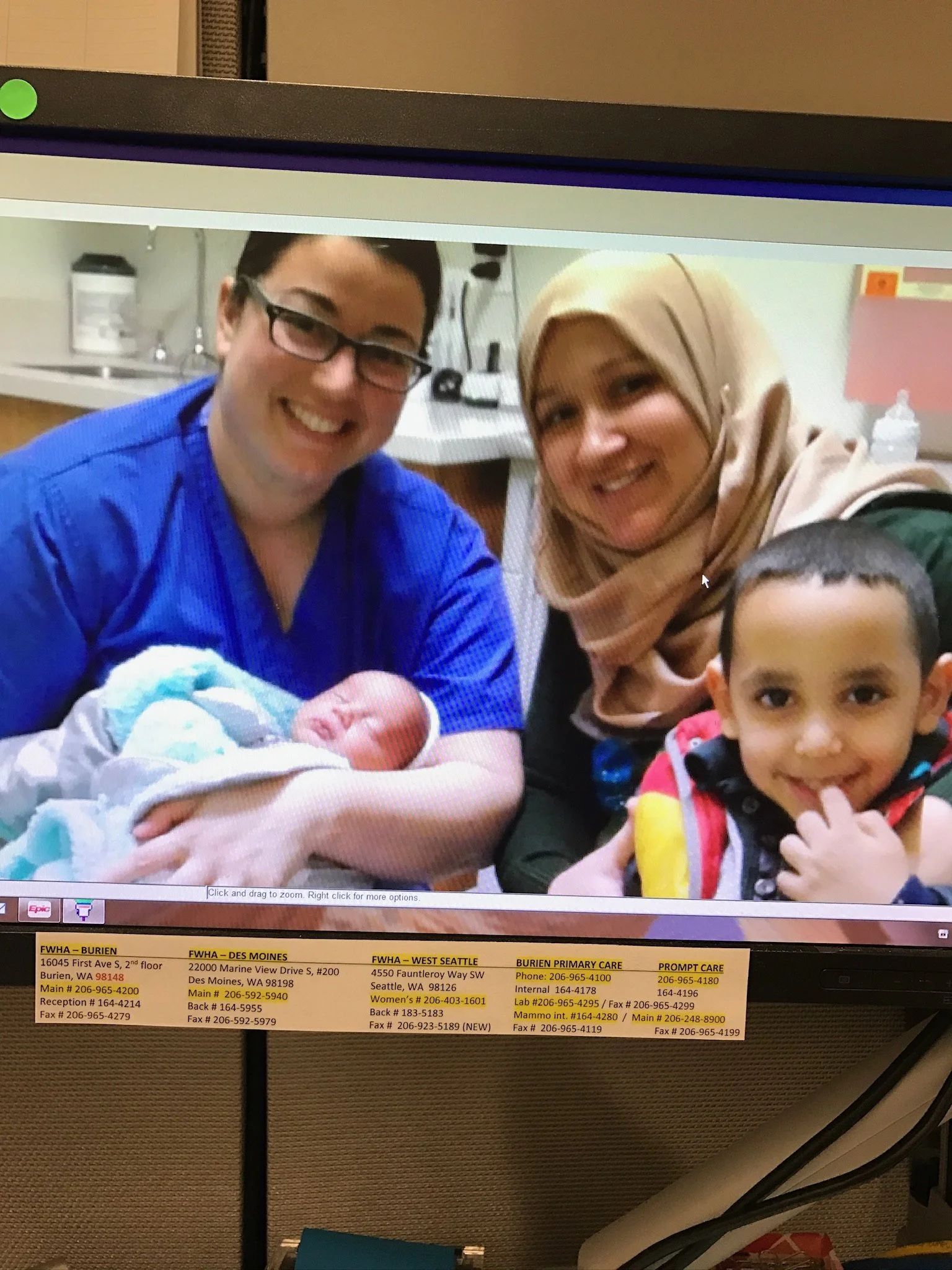 A smiling healthcare worker holding a newborn baby in a hospital room, with a woman wearing a hijab and a young boy smiling beside them.
