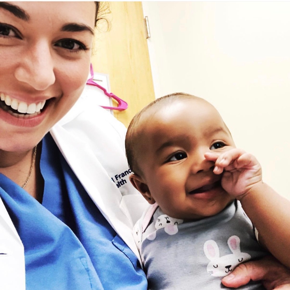 A smiling woman in medical scrubs holding a baby with a bunny print shirt, both smiling and looking happy.