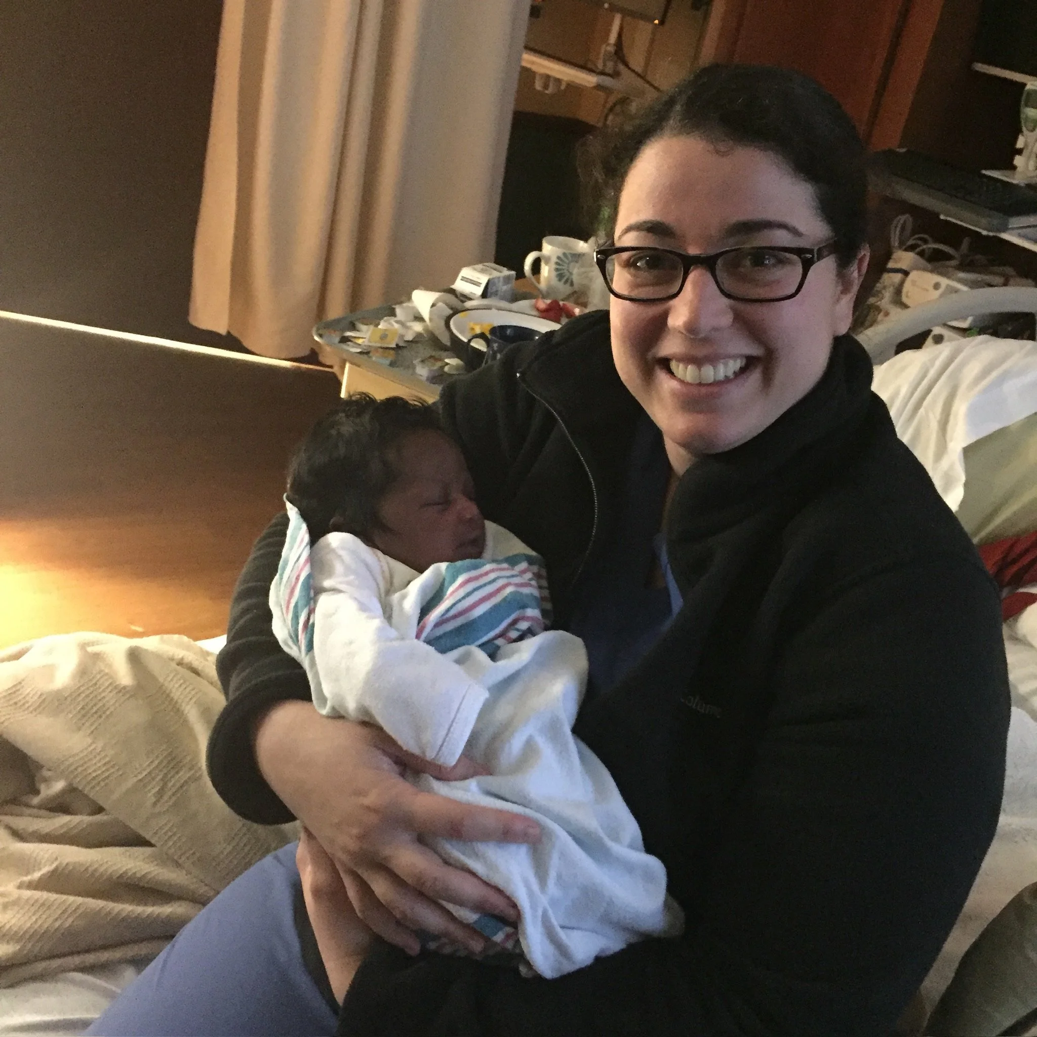 A woman in glasses and a black jacket is smiling while holding a newborn baby in a hospital room.