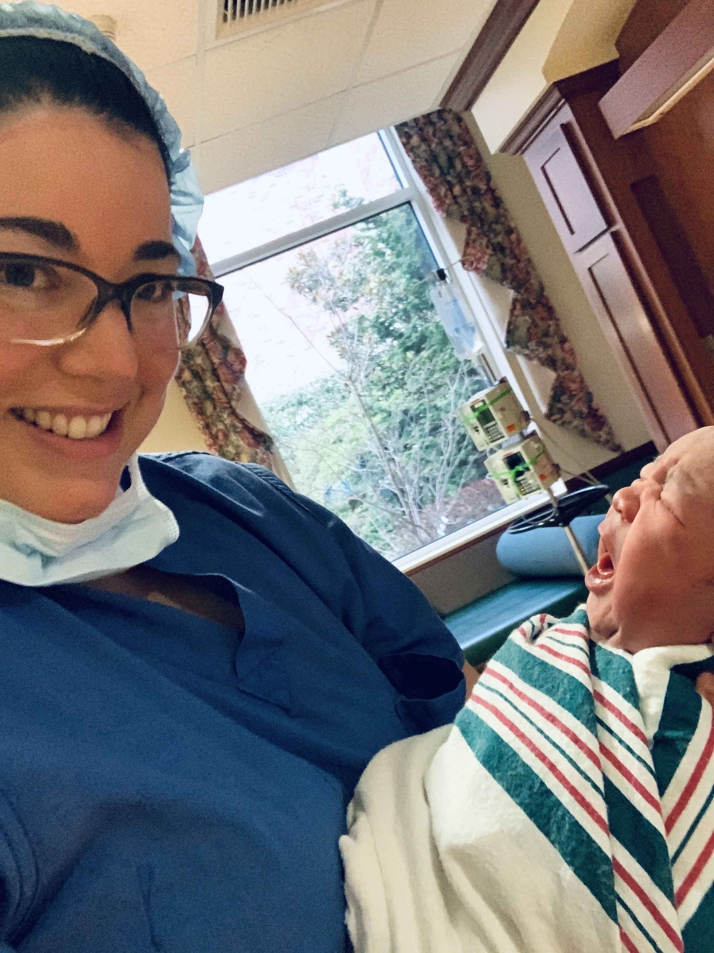 A smiling nurse in blue scrubs and glasses takes a selfie with a crying newborn baby in a hospital room. The baby is wrapped in a striped hospital blanket and is lying on a bed next to a window with floral curtains.