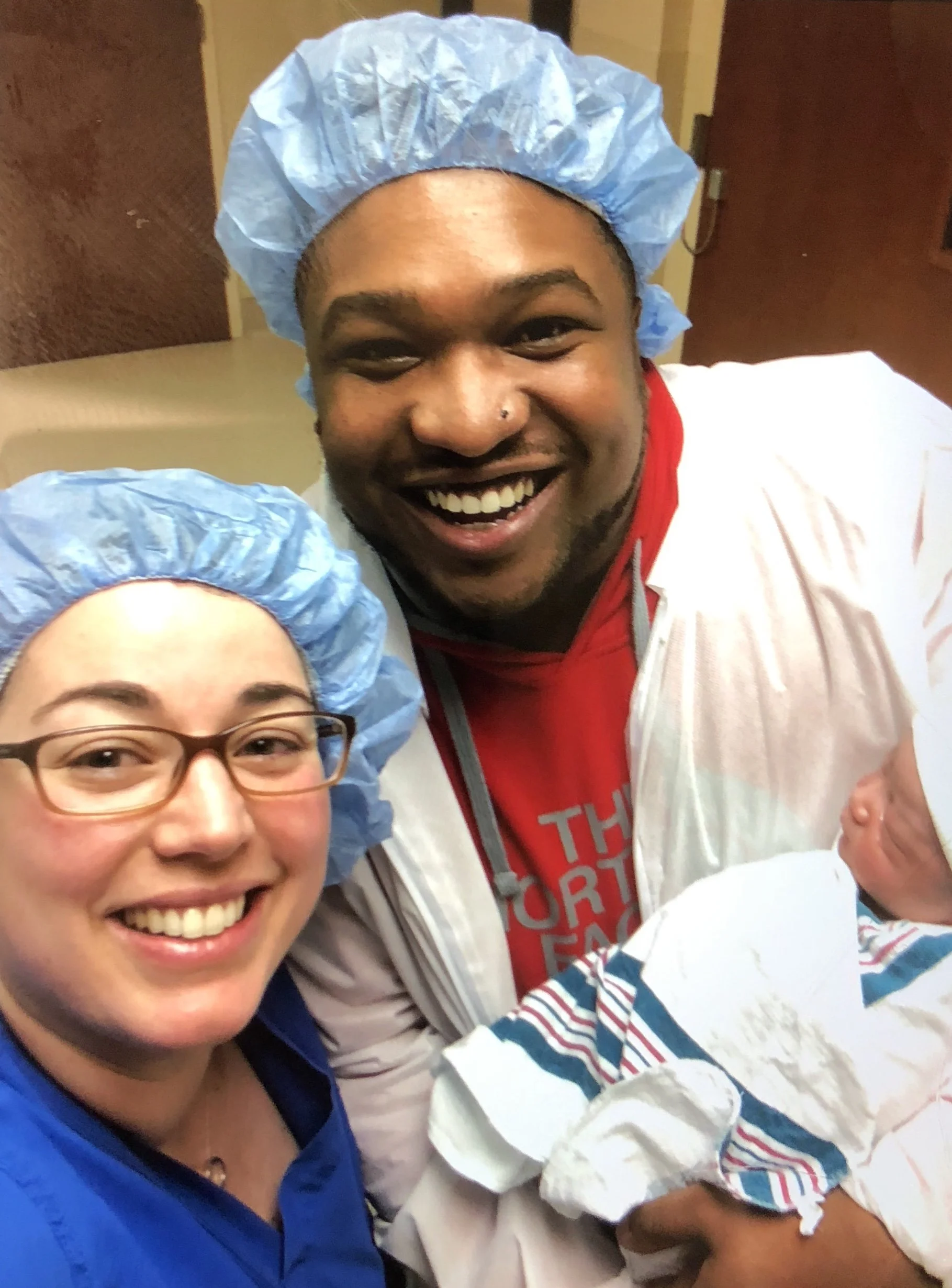 Happy couple wearing blue surgical caps, celebrating the birth of their baby in a hospital. The woman has glasses and is smiling, while the man is holding a newborn wrapped in a striped blanket.