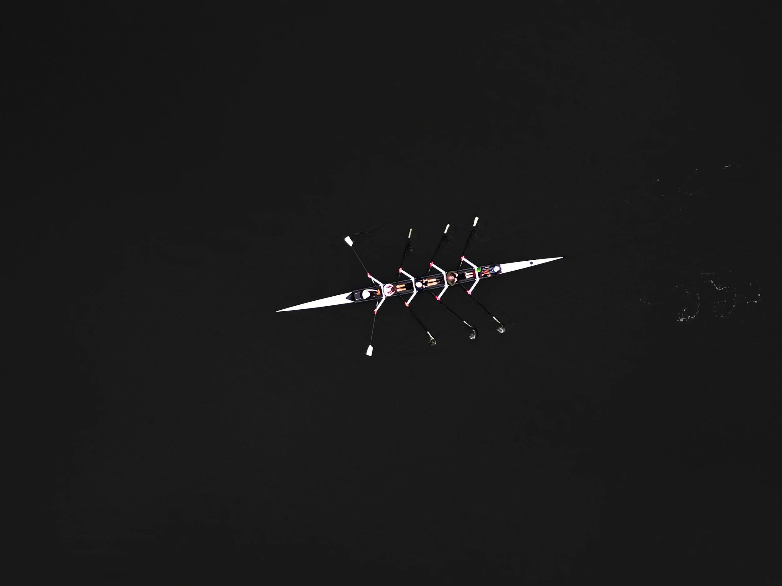 Representing Ananta Strategic's MISSION-FOCUSED EXECUTION, an aerial view of a rowing boat with four rowers on dark water, all rowing unison.