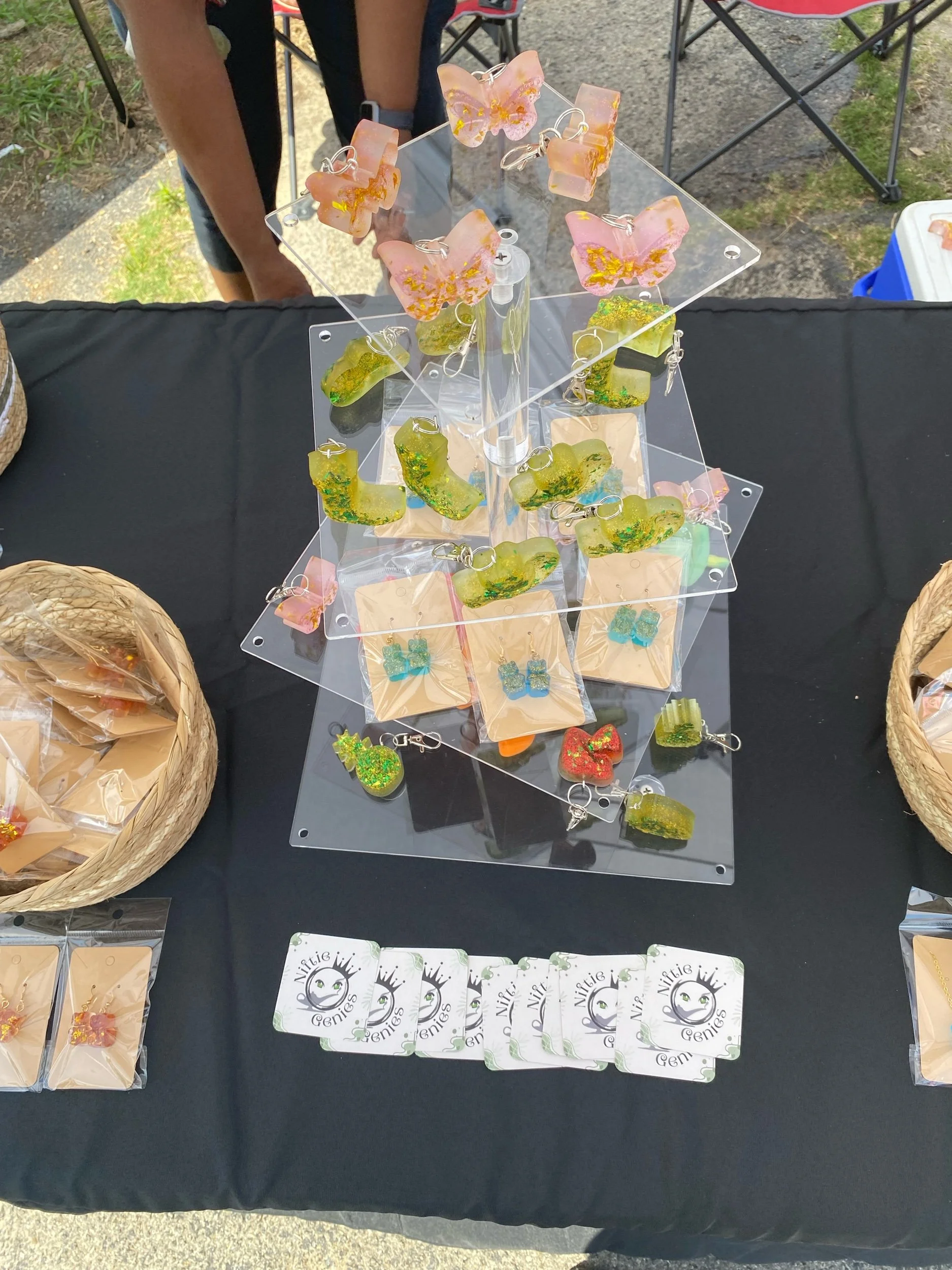Colorful resin keychains shaped like butterflies, bears, and other shapes displayed on a rotating stand at an outdoor market. There are business cards reading 'Niftie Genies' and baskets with necklaces and earrings on a black table.
