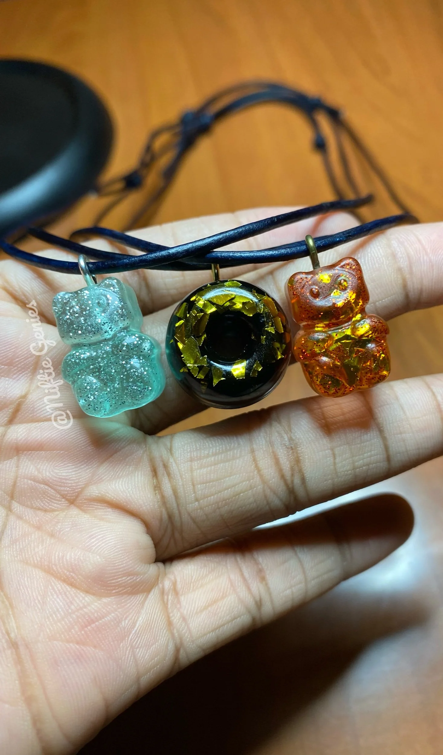 Close-up of a hand holding three small resin charms on a black cord, with a blurred wooden surface in the background.