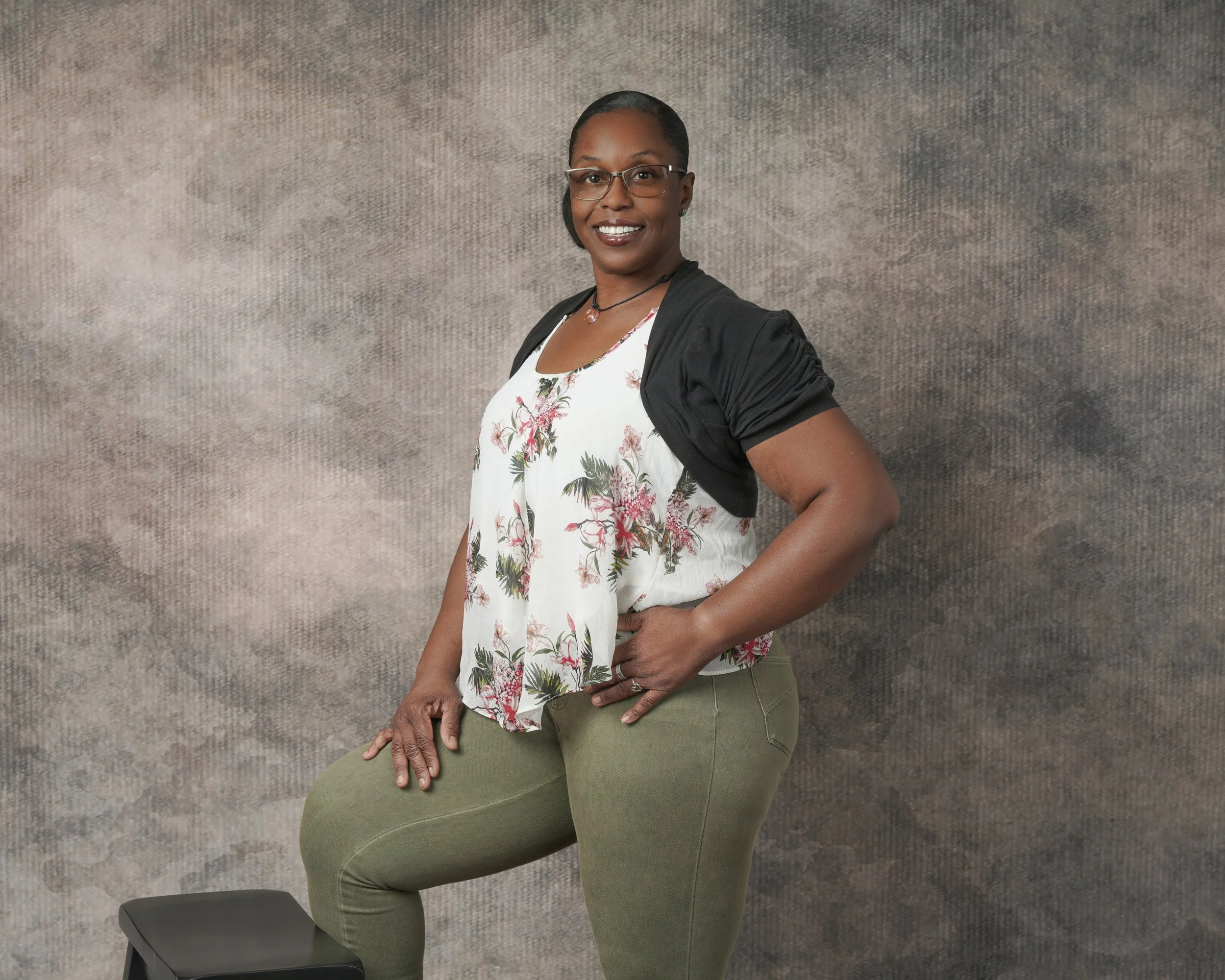 A confident woman with glasses and short black hair, smiling, dressed in a floral top with a black cardigan, green pants, and a necklace, standing in front of a textured gray background.