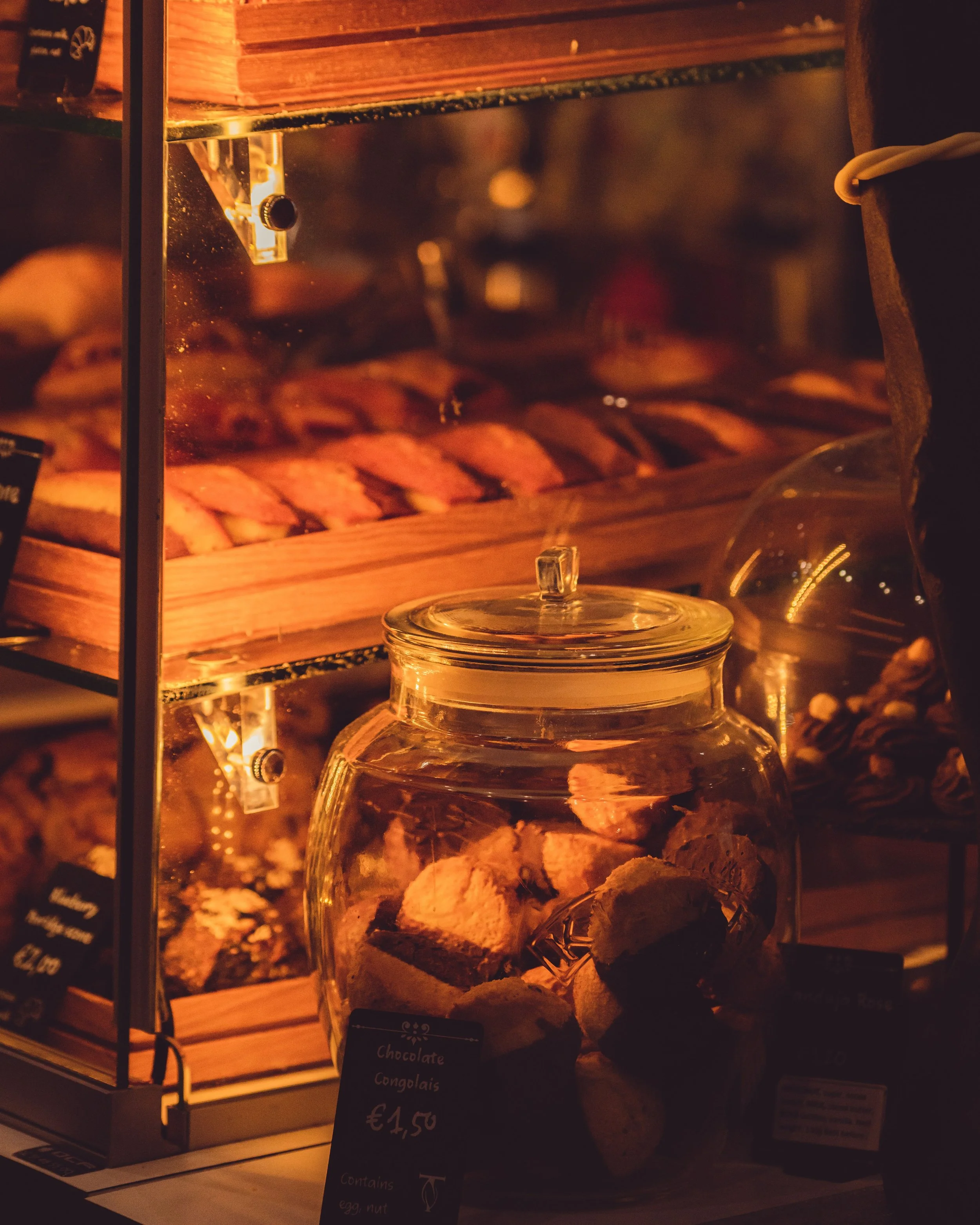 Glass display case with cookies inside, illuminated by warm light. Behind the case, there is a wooden wall.