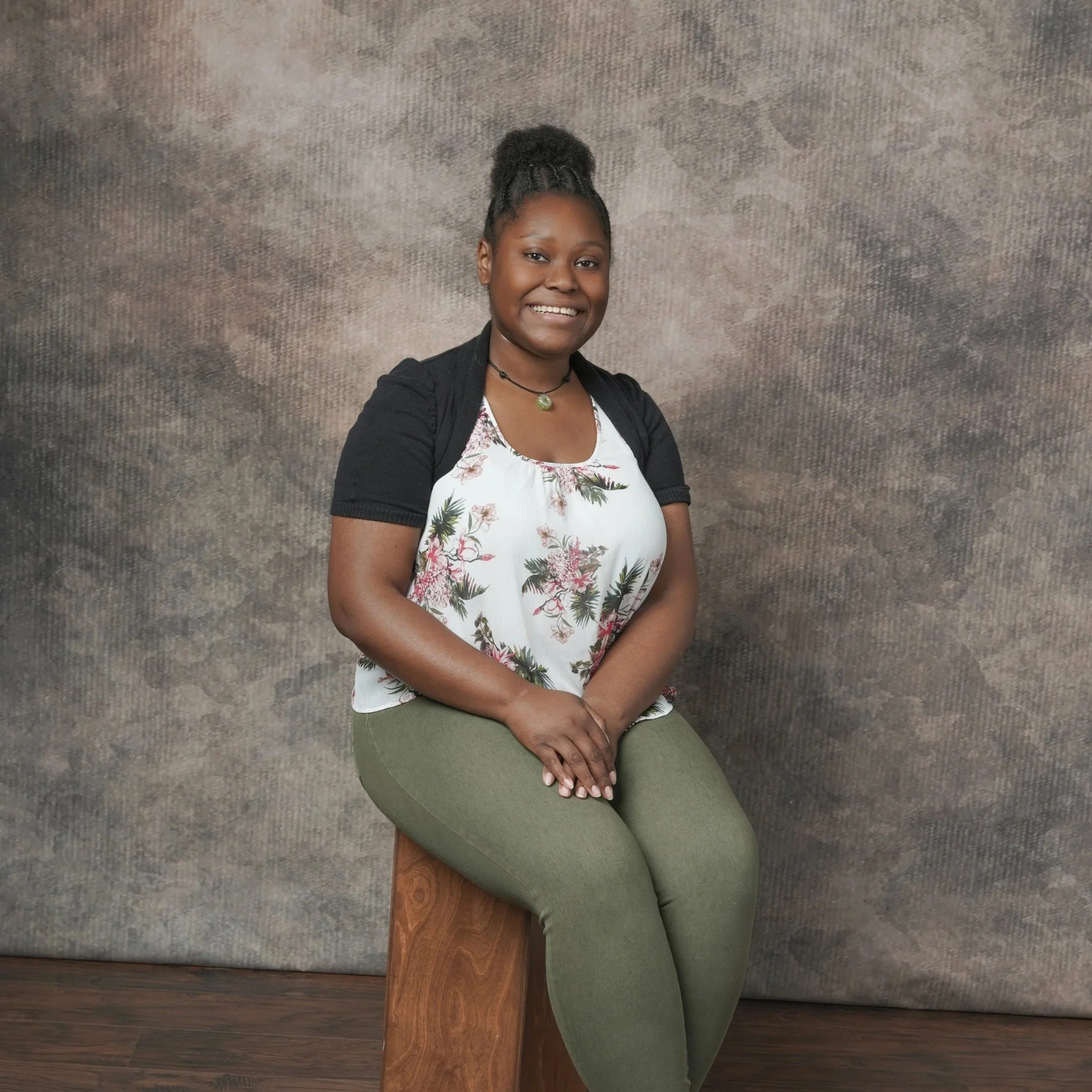 A woman with dark skin, dark braided hair in a bun, smiling, sitting with her hands on her lap against a textured wall. She wears a floral top, green pants, a black jacket, and a necklace with a green pendant.