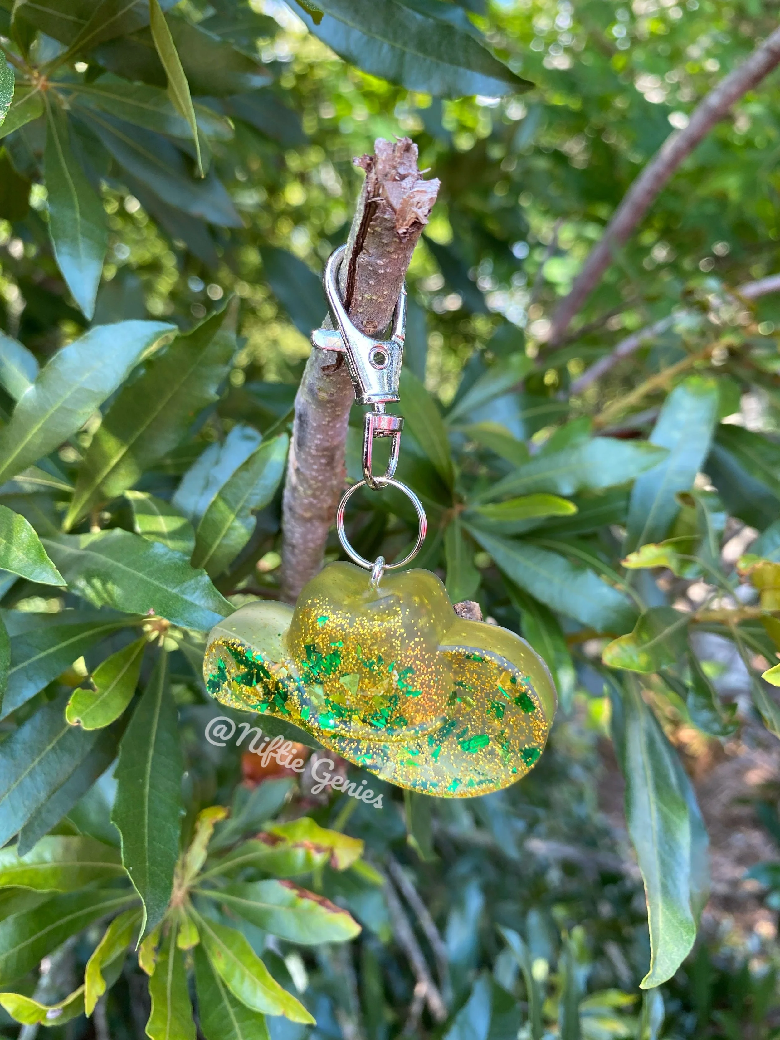 A close-up of a branch with green leaves and a glittery, yellow-green ornament hanging from a metal clip.