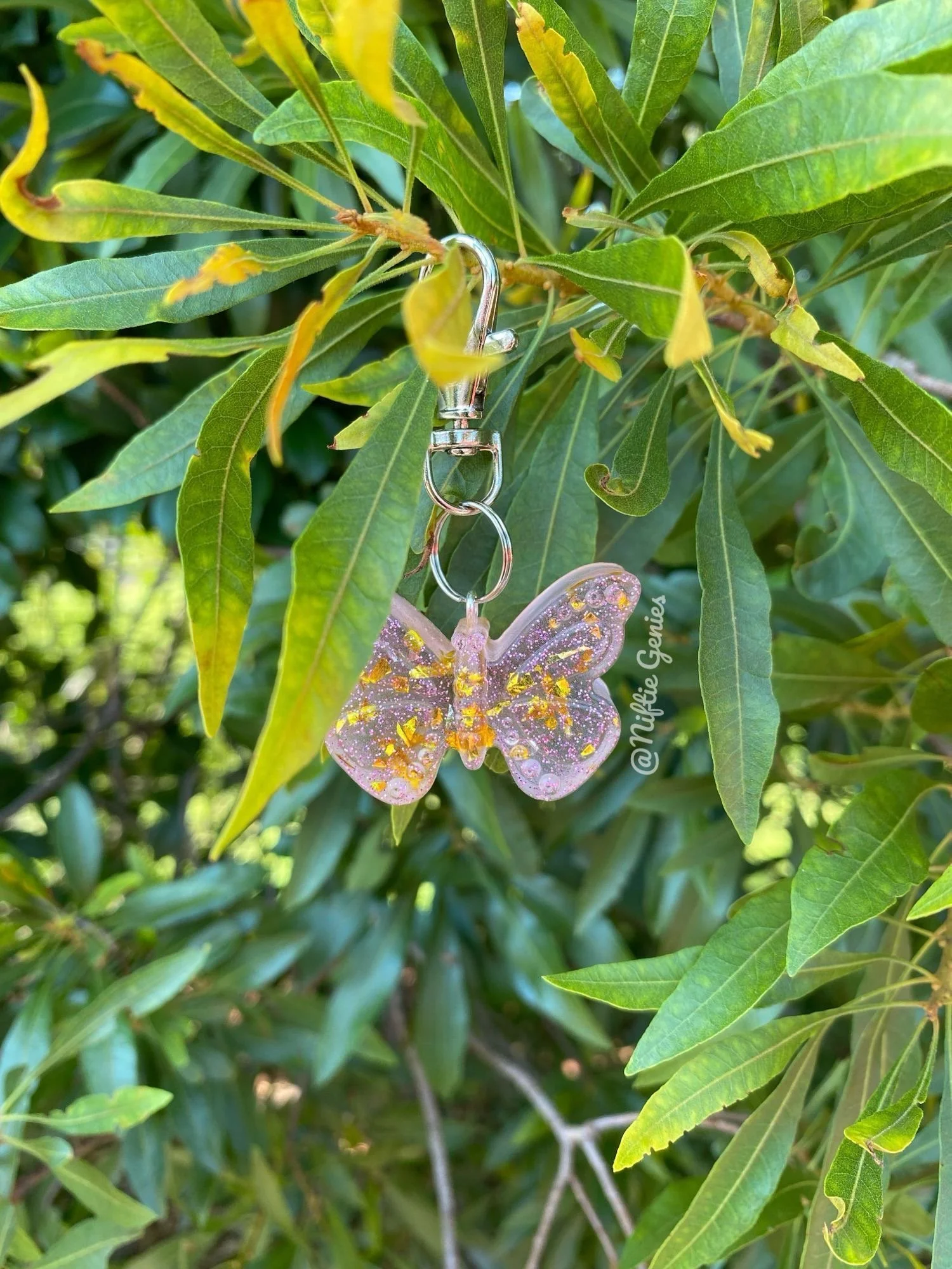 A close-up of a green leafy plant with a pink and glittery butterfly-shaped keychain hanging from one of the leaves.