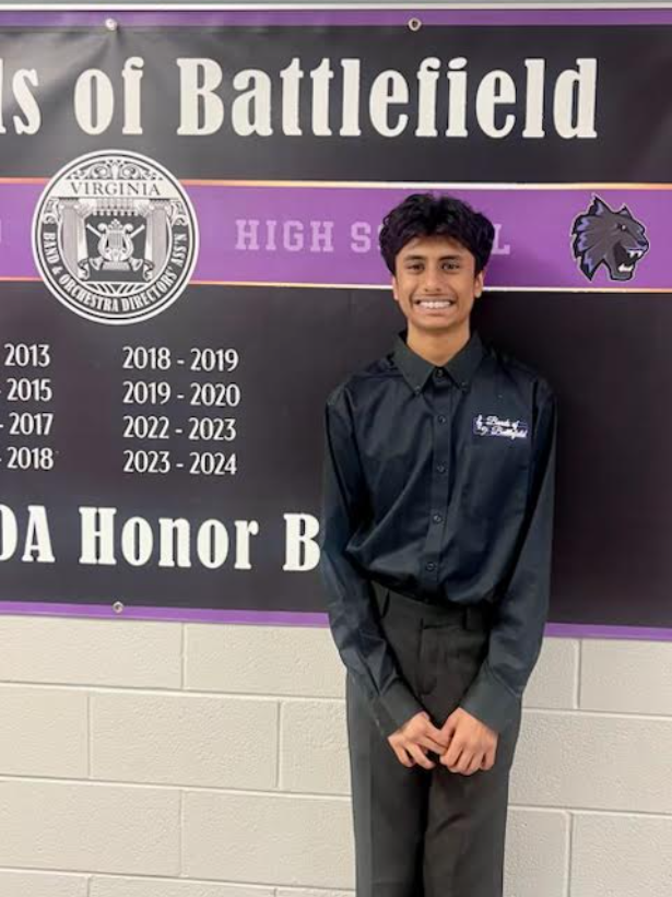A young man in a black button-up shirt and gray pants standing in front of a school banner with a purple and black design. The banner features the school’s seal, a wolf mascot, and lists academic years and honors. The young man is smiling, with short curly hair, and holds his hands together in front.