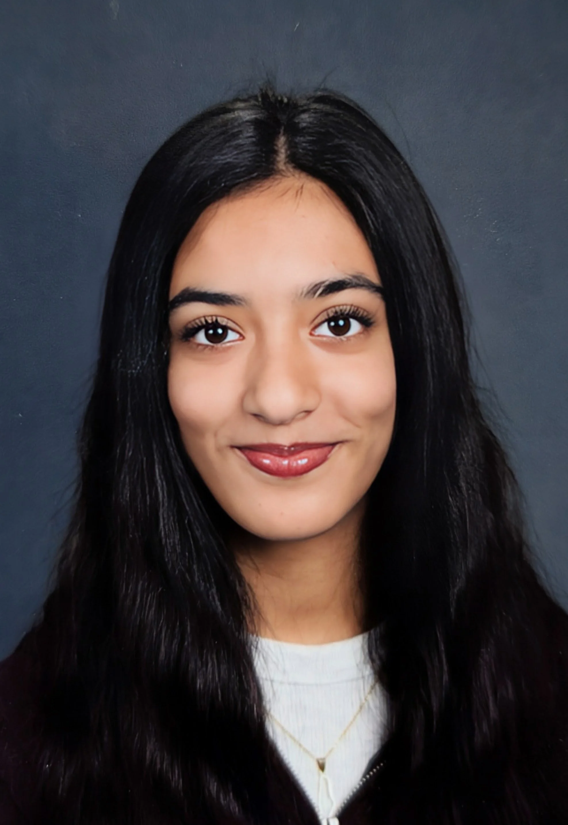 Close-up portrait of a young woman with long black hair, brown eyes, and a warm smile, wearing a white top, a black fur jacket, and a delicate gold necklace against a dark background.