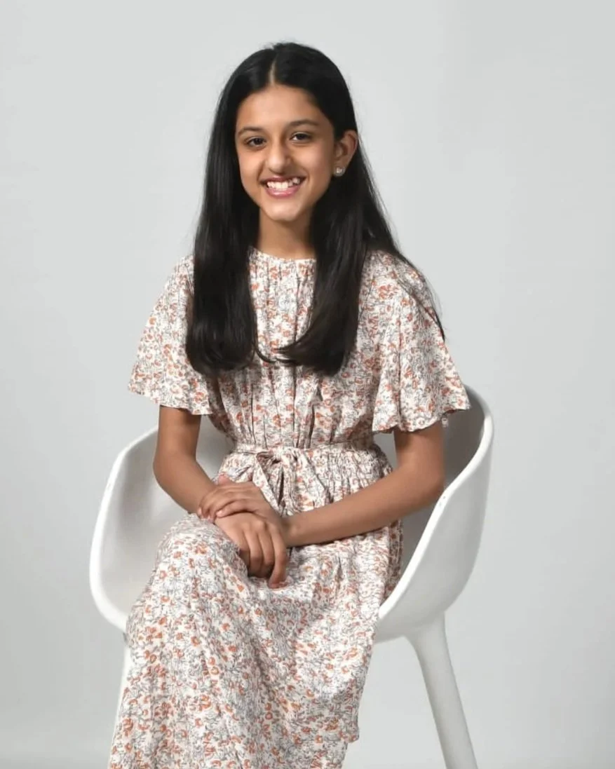 A young girl with long dark hair wearing a floral dress sitting on a white chair against a plain light grey background.
