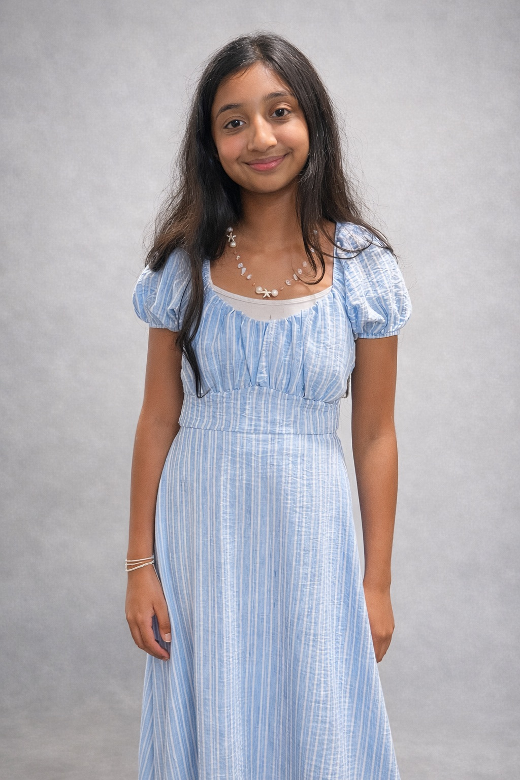 A young girl with long dark hair wearing a light blue striped dress, a shell necklace, and a bracelet, standing against a plain grey background and smiling softly.