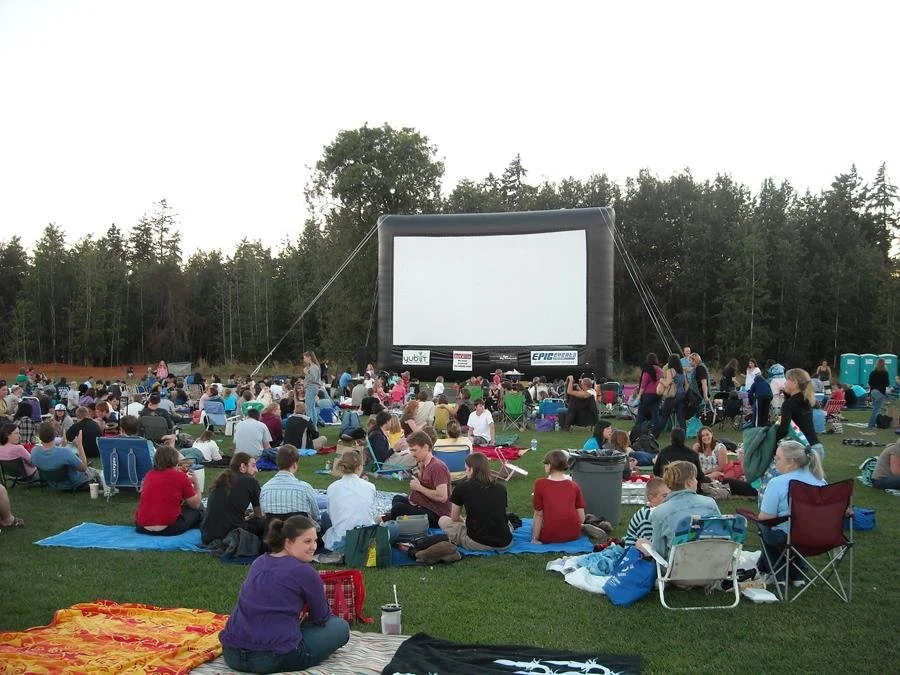 Outdoor outdoor movie screening at dusk with a crowd sitting on blankets and chairs in front of a large inflatable movie screen, surrounded by trees.