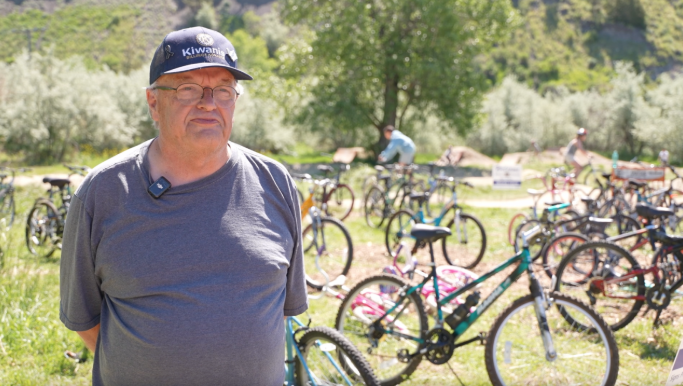 Older man in gray t-shirt and cap standing outdoors among numerous bicycles on a grassy field, with trees in the background.