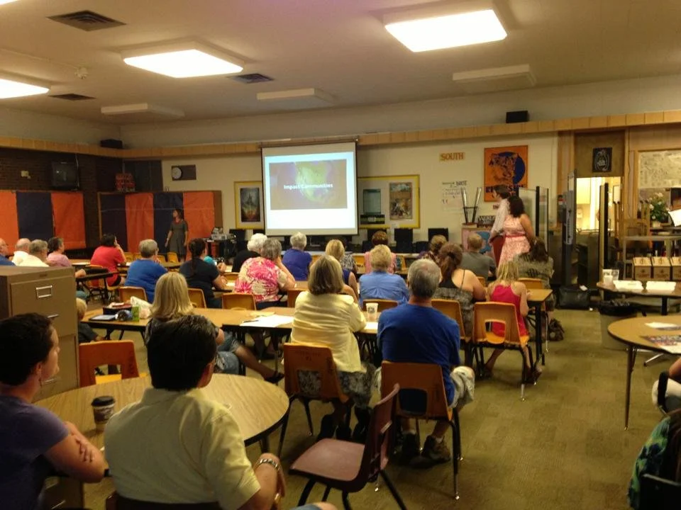Group of adults attending a presentation in a classroom or community center, with a woman speaking at the front and a projector screen displaying a slide.