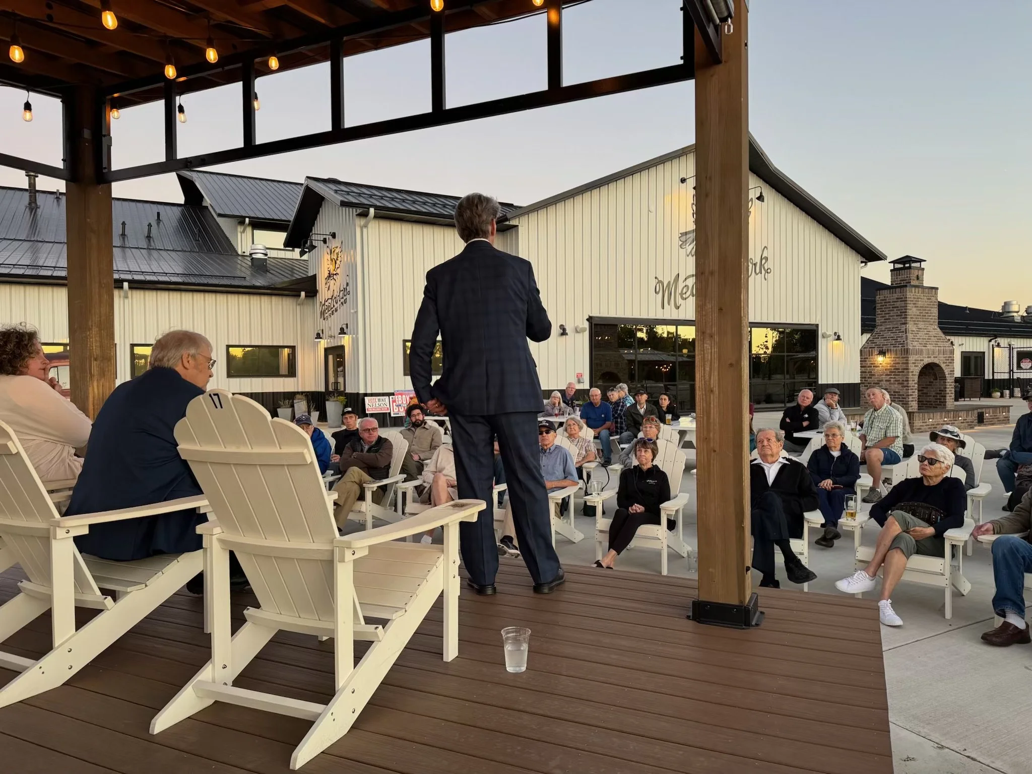 A man in a dark suit speaking to an outdoor audience seated on chairs. The event is taking place on a patio with wooden flooring and a roof with string lights. In the background, there's a modern building with large windows and an outdoor fireplace.