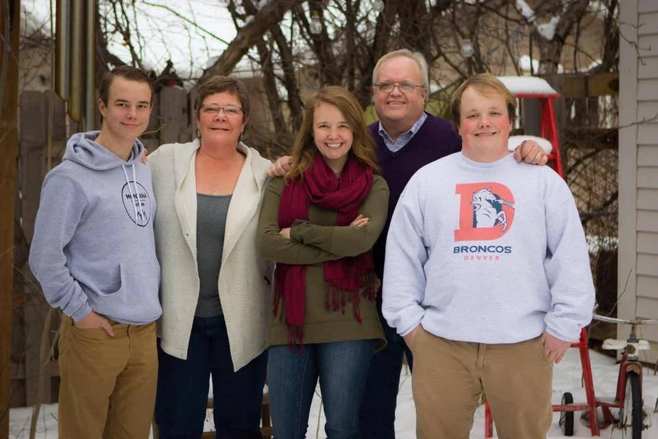 A family of five standing outdoors in a backyard with trees and a fence, posing for a photo during winter. The family includes two teenagers, two adults, and a younger boy, all smiling and dressed warmly.