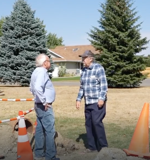 Two men conversing outdoors near orange traffic cones and dirt patch, with evergreen trees and a house in background.