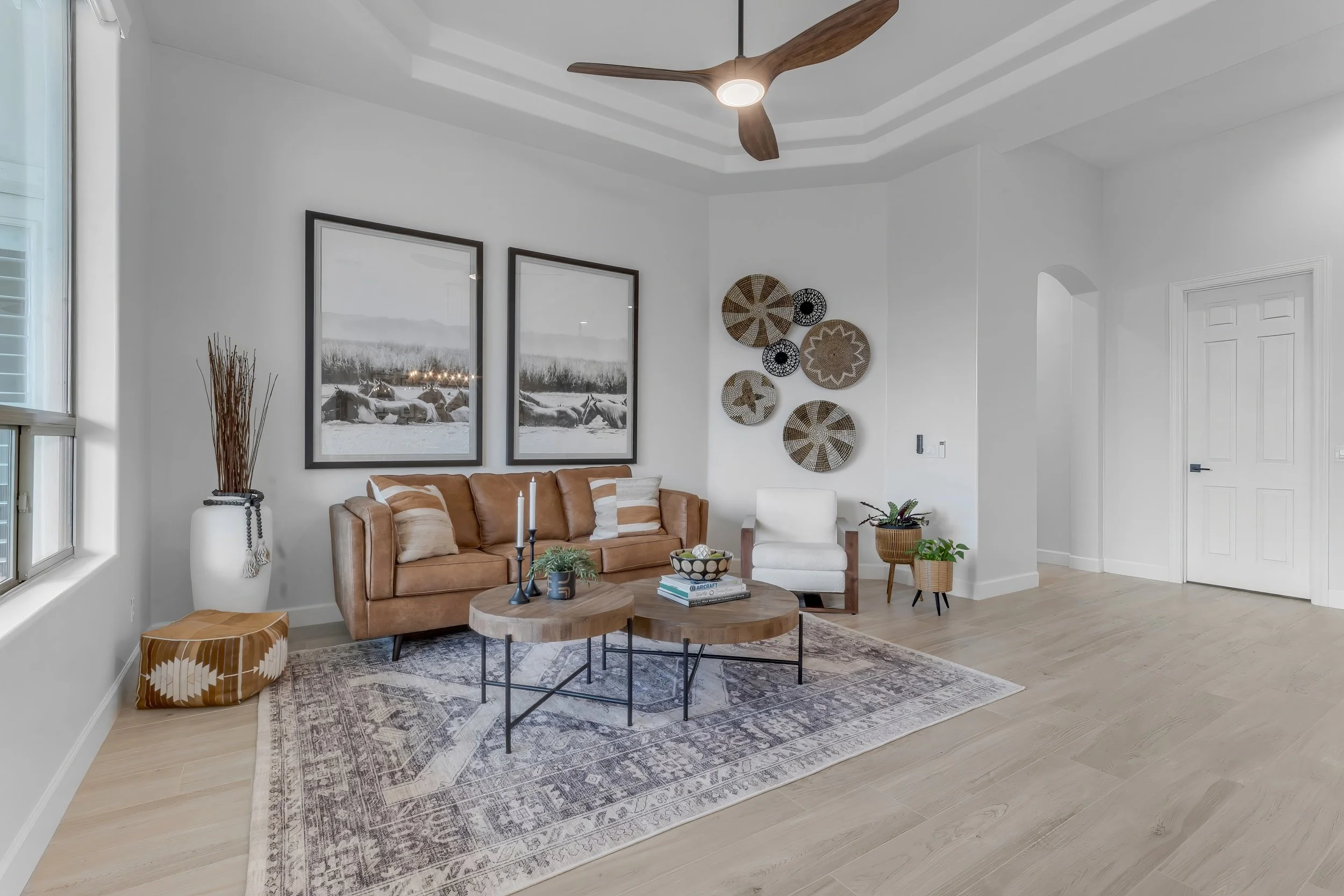 Bright living room with white walls, a leather sofa, two round wooden coffee tables, a white armchair, and wall art including framed black-and-white landscape photos and woven baskets. The room has a light hardwood floor and a ceiling fan.