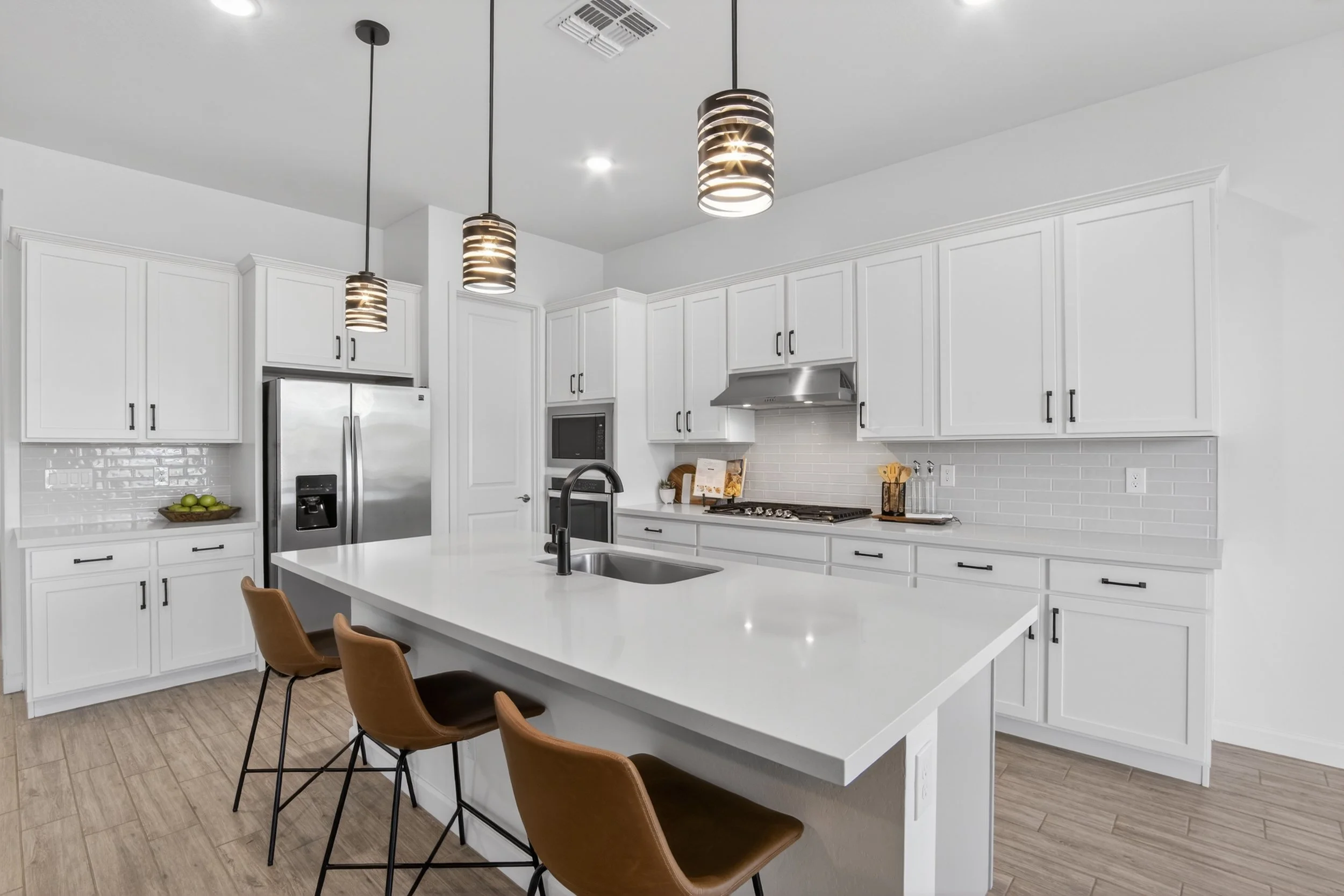 Modern kitchen with white cabinets, a large white island with a sink, three brown chairs, stainless steel appliances, pendant lights, and a wooden floor.