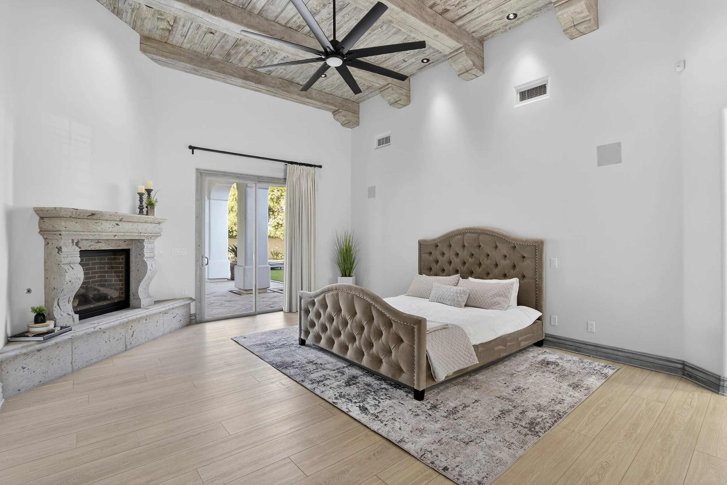 Elegant bedroom with a high ceiling featuring exposed wooden beams and a modern black ceiling fan. Light-colored hardwood flooring, a large upholstered bed with tufted headboard and multiple pillows, a vintage fireplace, and sliding glass doors leadi