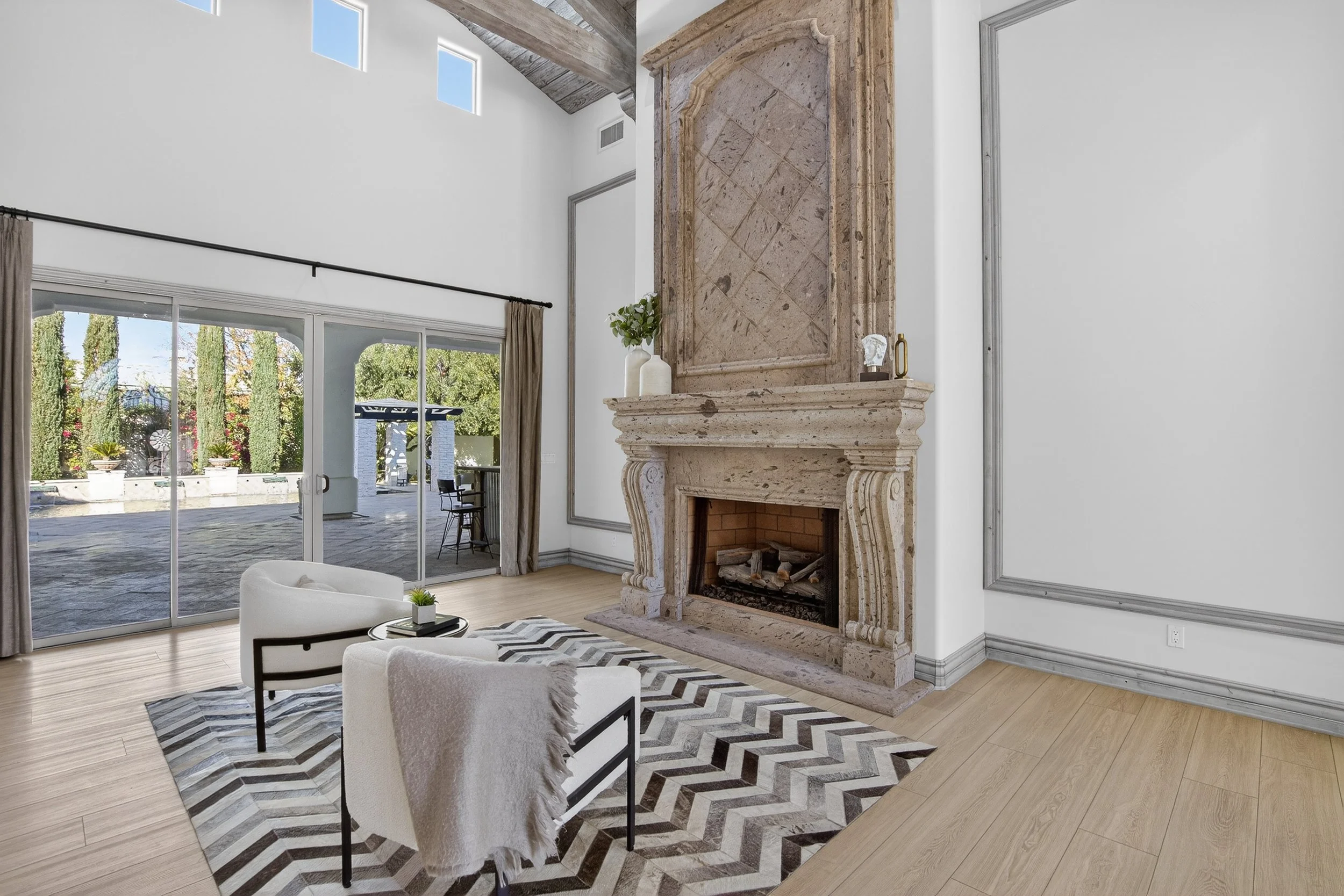 Living room with a large stone fireplace, sliding glass doors leading to an outdoor patio, modern furniture including a white armchair and a black and white chevron-patterned rug, and a view of a landscaped backyard with tall trees.