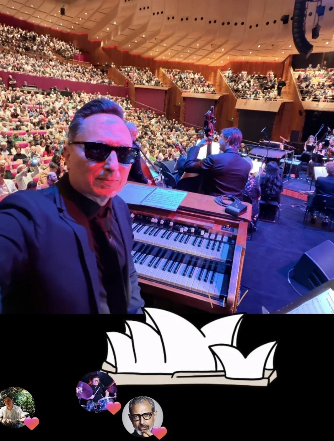 A man in sunglasses taking a selfie  on stage at The Sydney Opera House with an orchestra performing on stage and a large audience in the background.