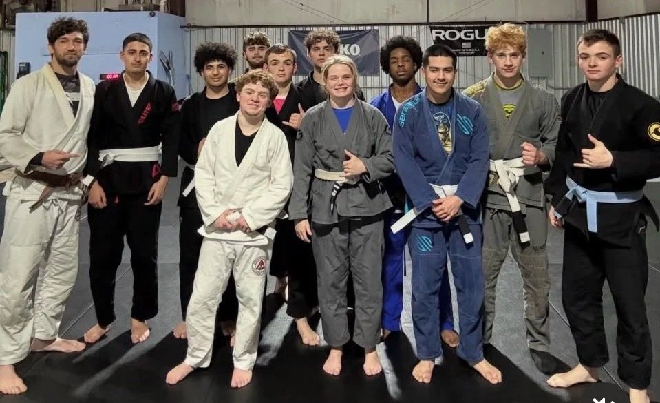 Group of young athletes in Brazilian Jiu-Jitsu gis and belts, standing in a training gym, smiling for a photo.