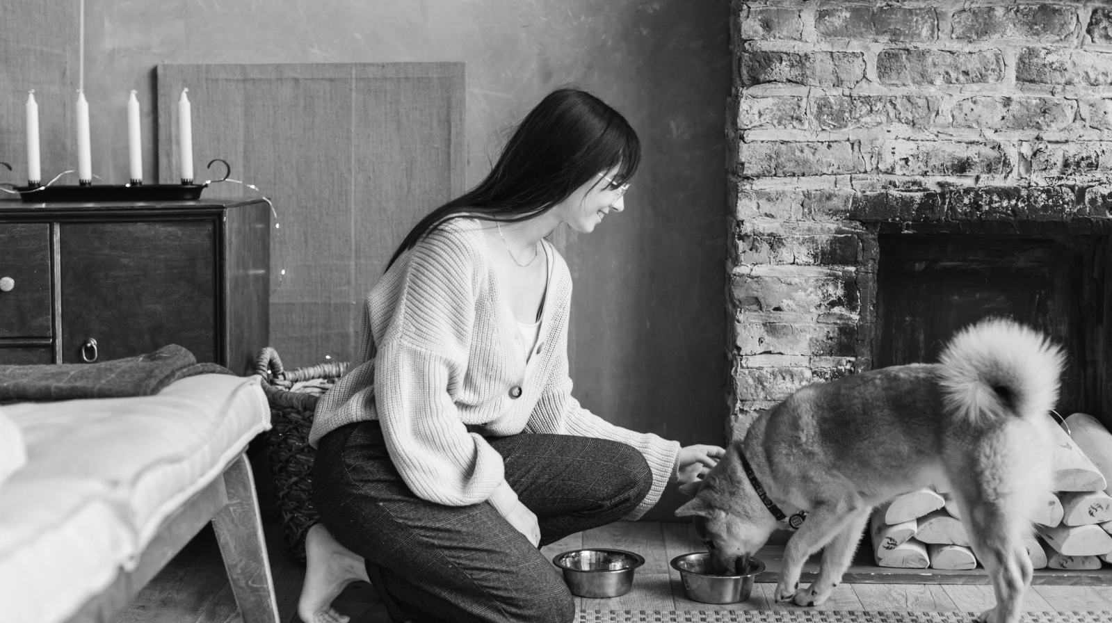 A woman with long dark hair sitting on the floor near a fireplace, feeding a dog from a bowl. The scene is in a cozy room with a brick fireplace, candles on a cabinet, and a pile of logs.
