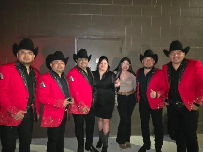 Group of six people, five men in red and black cowboy outfits with black hats and two women in black and gray outfits, standing together in front of a gray wall. They are smiling and posing for the photo.