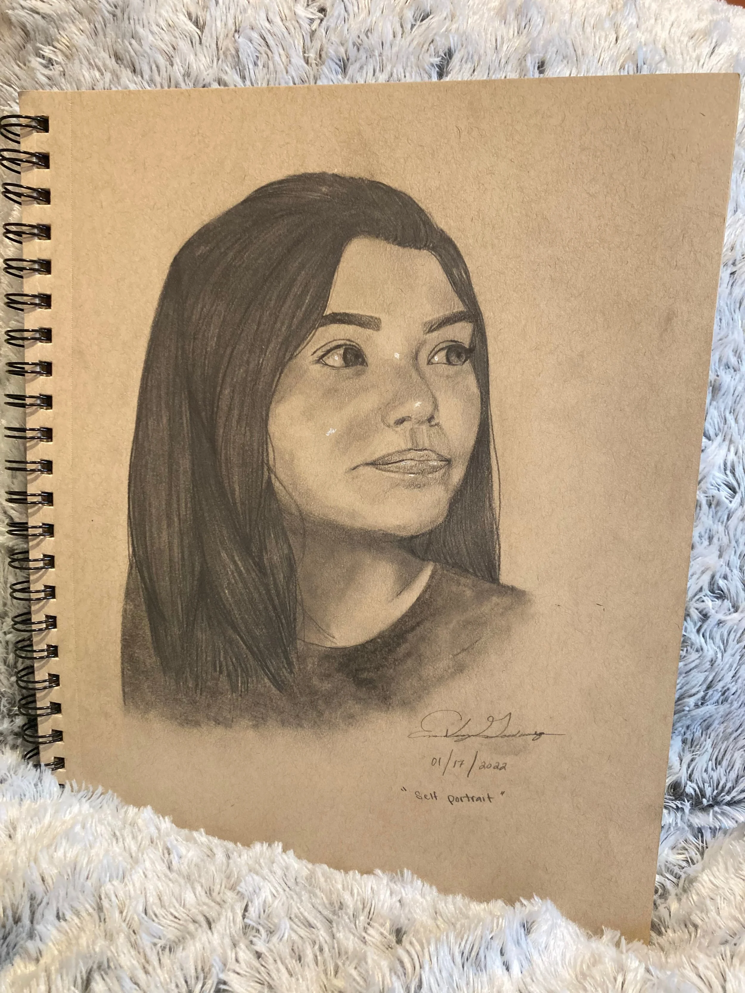 A detailed pencil sketch of a woman with long hair on beige paper. The drawing is part of a spiral-bound sketchbook placed on a white fluffy surface. The bottom right of the page has handwritten text with the date 01/17/2022 and the title 'Self Portr