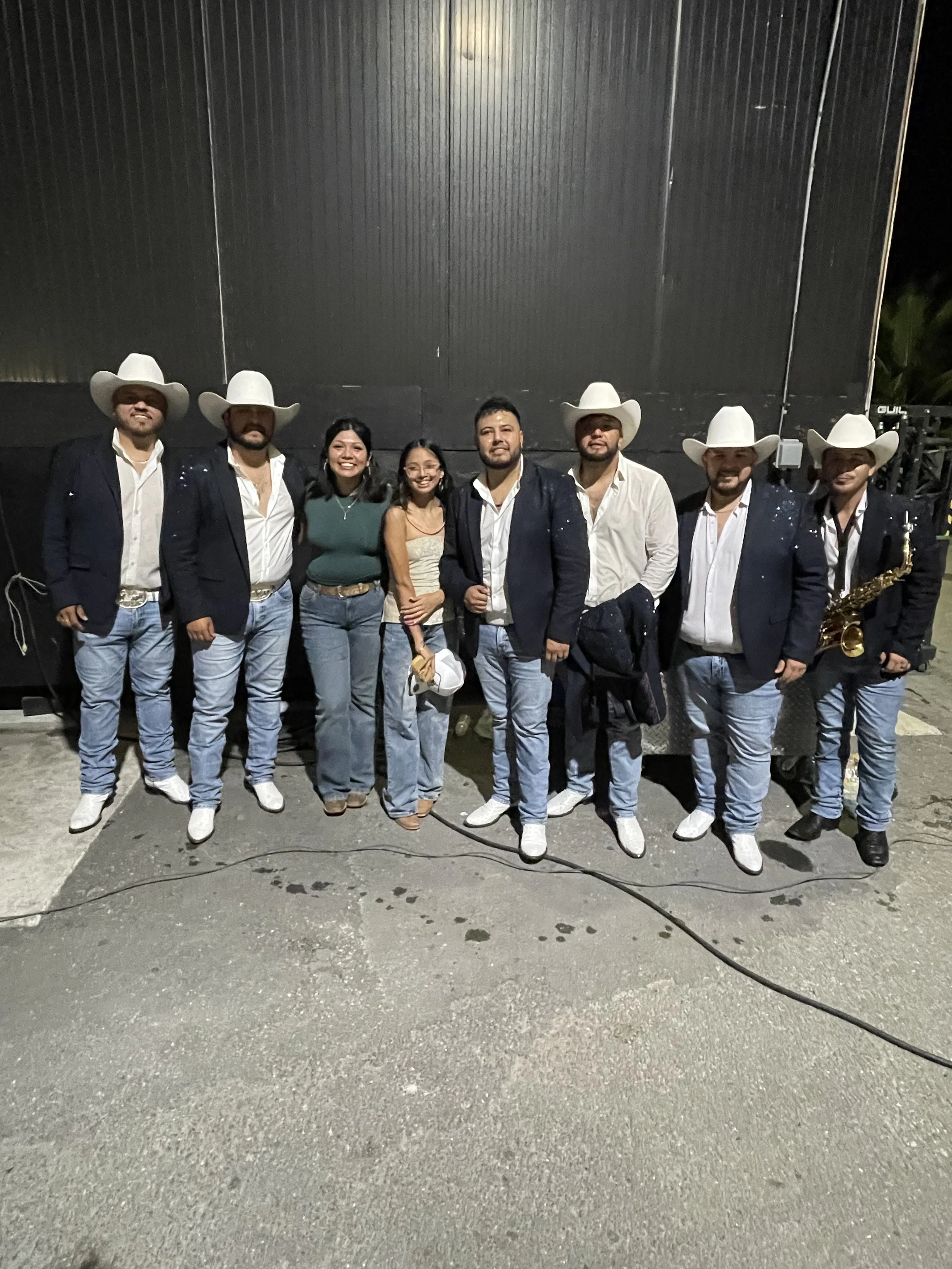 A group of nine people standing together outdoors at night, dressed in formal and Western-style clothing, some wearing cowboy hats, with a large black wall in the background.