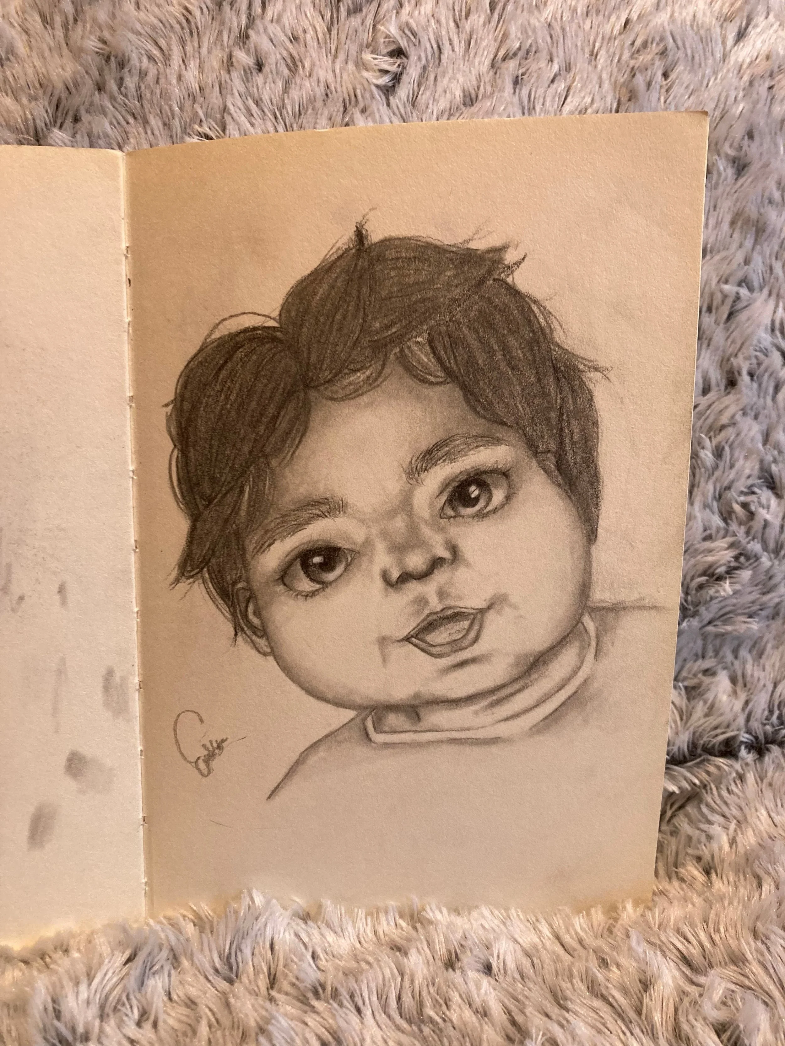 A detailed pencil drawing of a young child's face showing expressive eyes, full cheeks, and soft lips, with tousled hair, on a piece of paper placed on a plush, textured surface.