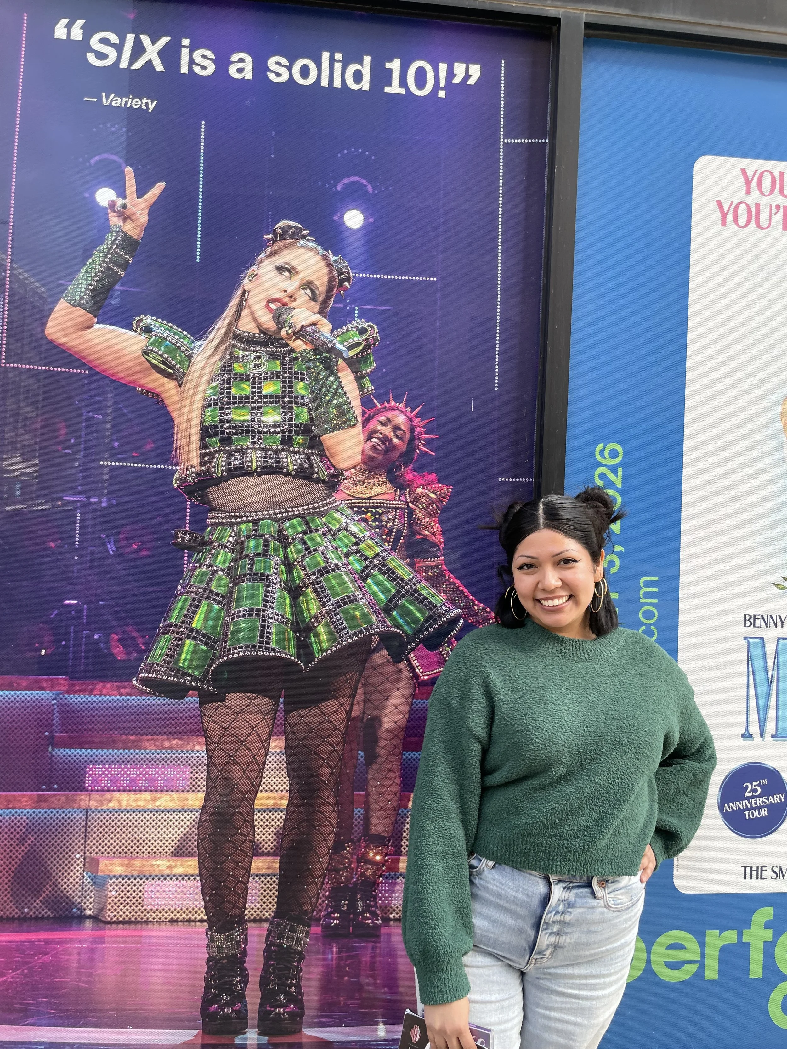 A young woman with black hair, wearing a green sweater and light blue jeans, smiling and standing in front of a large poster of the broadway poster for the musical Six.