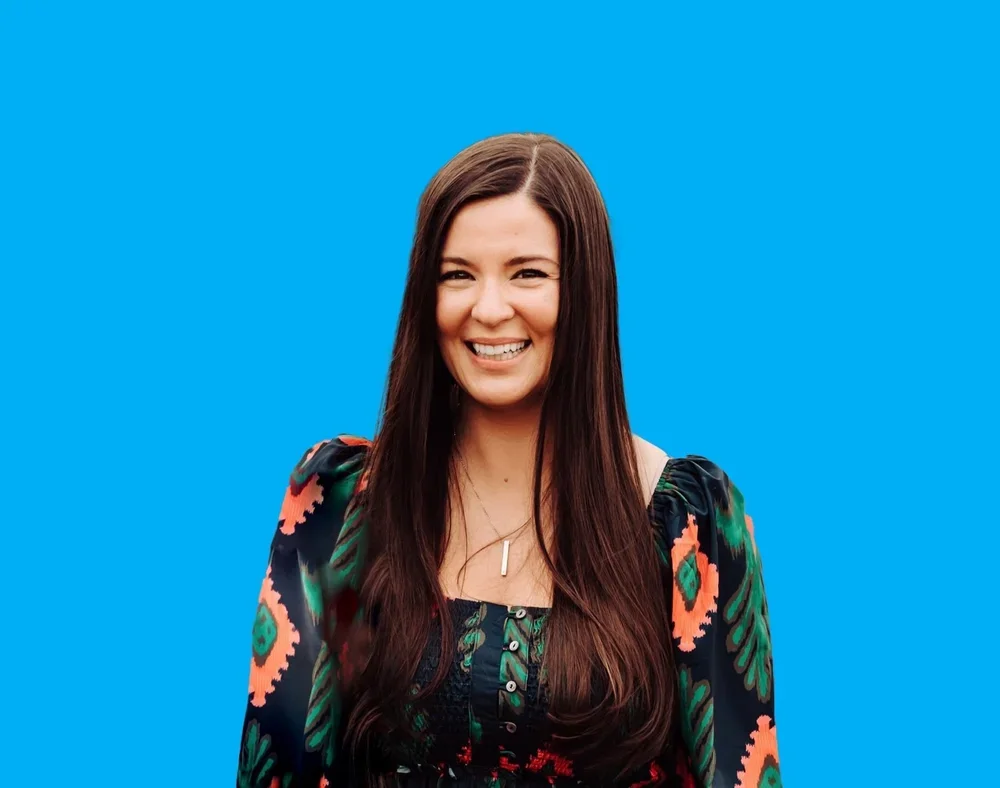 A woman with long brown hair smiling against a plain blue background, wearing a black top with colorful floral and abstract patterns.