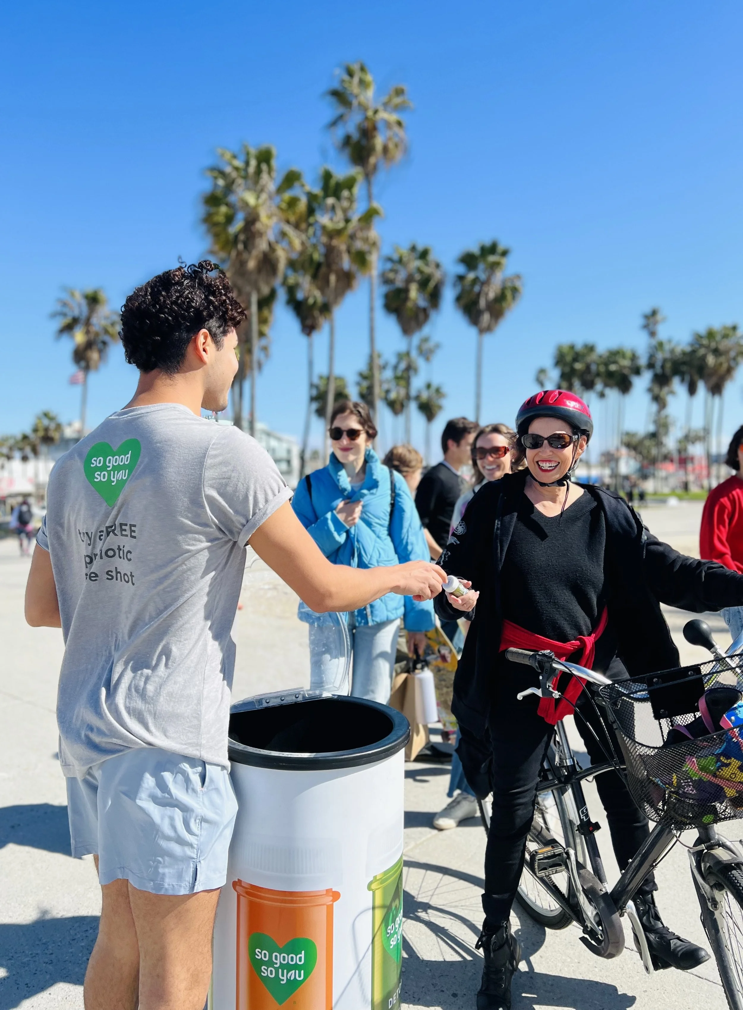 A man in a gray t-shirt and shorts is handing out bottles of water to a smiling woman wearing a helmet and sunglasses at a beachside location. Several people are in the background, with palm trees and a clear blue sky overhead.
